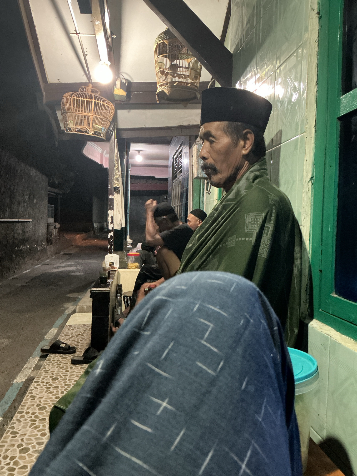 The image depicts an outdoor scene at night, likely on a street or alley in Magelang, Indonesia. The foreground is dominated by the shoulder and lap of a person wearing a dark blue garment with a subtle white geometric pattern. To the right, a light green tiled wall with a green-framed window is visible, along with a light blue lidded container.
The middle ground features an older man with a mustache, wearing a black peci (a traditional cap) and a dark green patterned shawl draped over his shoulders. He is looking to his left. Behind him, other individuals are seated, some wearing similar peci hats. A birdcage hangs from the ceiling in the upper left portion of the image, and another is visible further back. The environment appears to be a covered walkway or porch with wooden beams and exposed lighting fixtures. In the background, a dark, narrow street or alleyway is visible, suggesting a residential or commercial area. The ground is paved with mosaic-like tiles. The overall impression is one of a casual gathering in the evening.