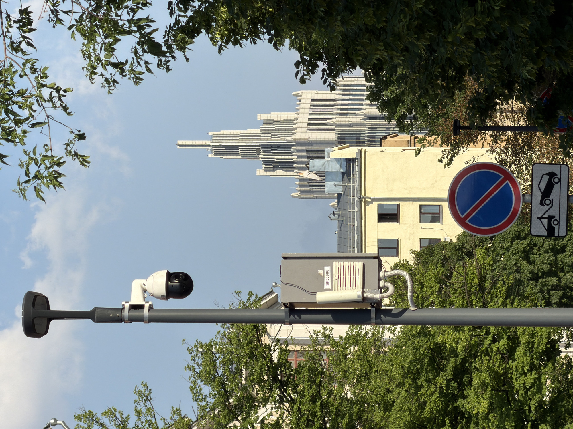 The image captures an urban scene in Moscow, Russia. The foreground features a gray utility pole with attached surveillance equipment, including a security camera and a gray electrical box labeled "SF50096." Behind the pole and its equipment, there are street signs, one a blue and red circle with a diagonal bar, signifying a "no stopping" zone, and another depicts a vehicle being towed. A cream-colored building is partially visible behind the signs. In the background, a modern, stepped skyscraper rises into the sky, flanked by treetops on both sides. The sky is light blue with scattered clouds.