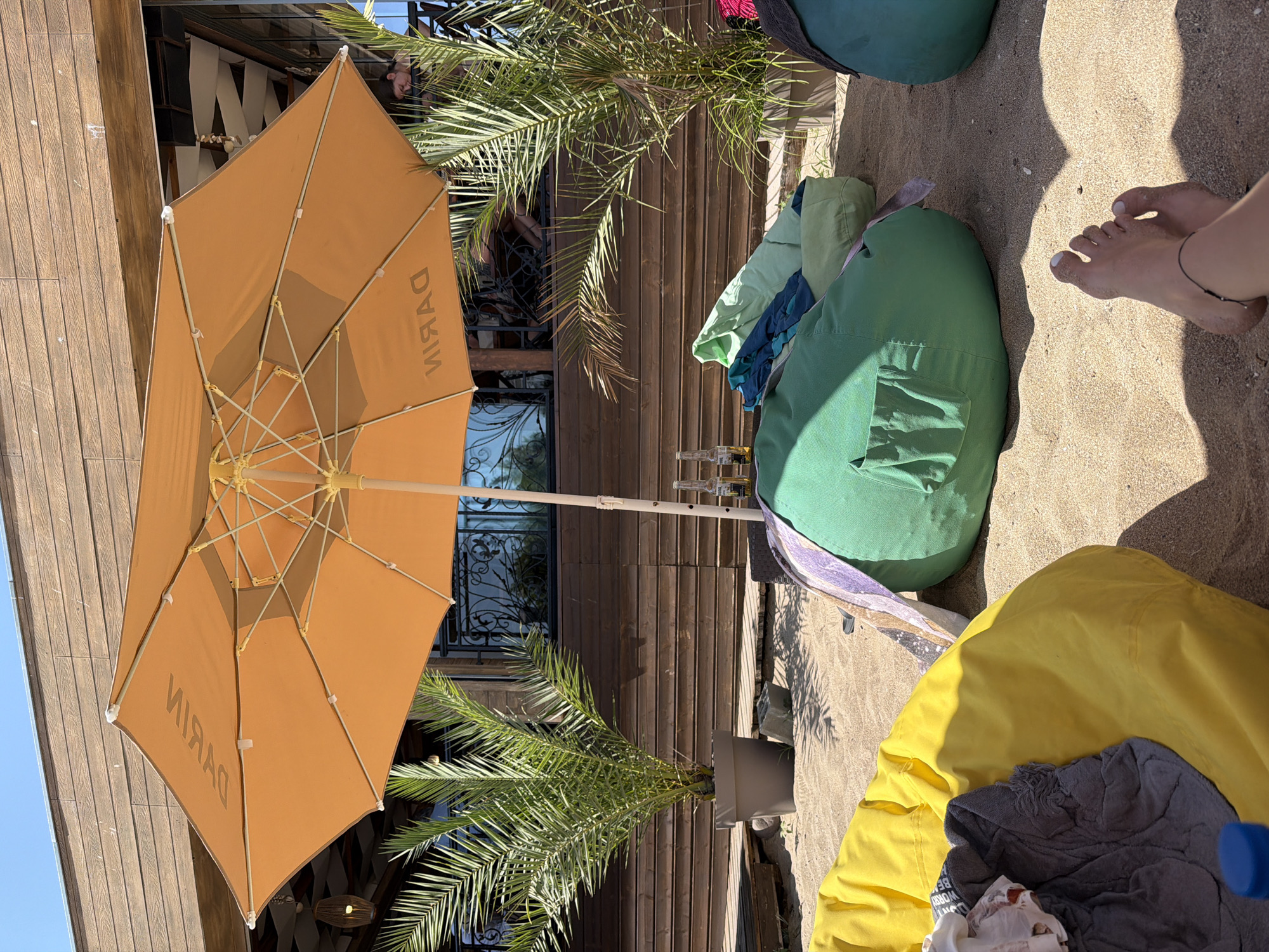 The image depicts a beachside seating area under a large orange umbrella. The umbrella has the word "DYKIM" written on it in reverse, suggesting it may be a brand or establishment name. In the foreground, there are several large beanbag chairs, one yellow and two green, scattered on the sand. A person's bare feet, with a black anklet, are visible in the lower right corner, indicating someone is relaxing. Two bottles of Corona beer stand on the sand near the umbrella's pole. Palm trees in large pots flank the seating area, and a wooden structure forms the backdrop, possibly part of a beach bar or restaurant. The overall scene suggests a casual, outdoor recreational space, likely a beach club or cafe, possibly located in Sozopol, Bulgaria, given the beach setting and common presence of such establishments in tourist areas. The lighting indicates it is daytime, with shadows cast on the sand.