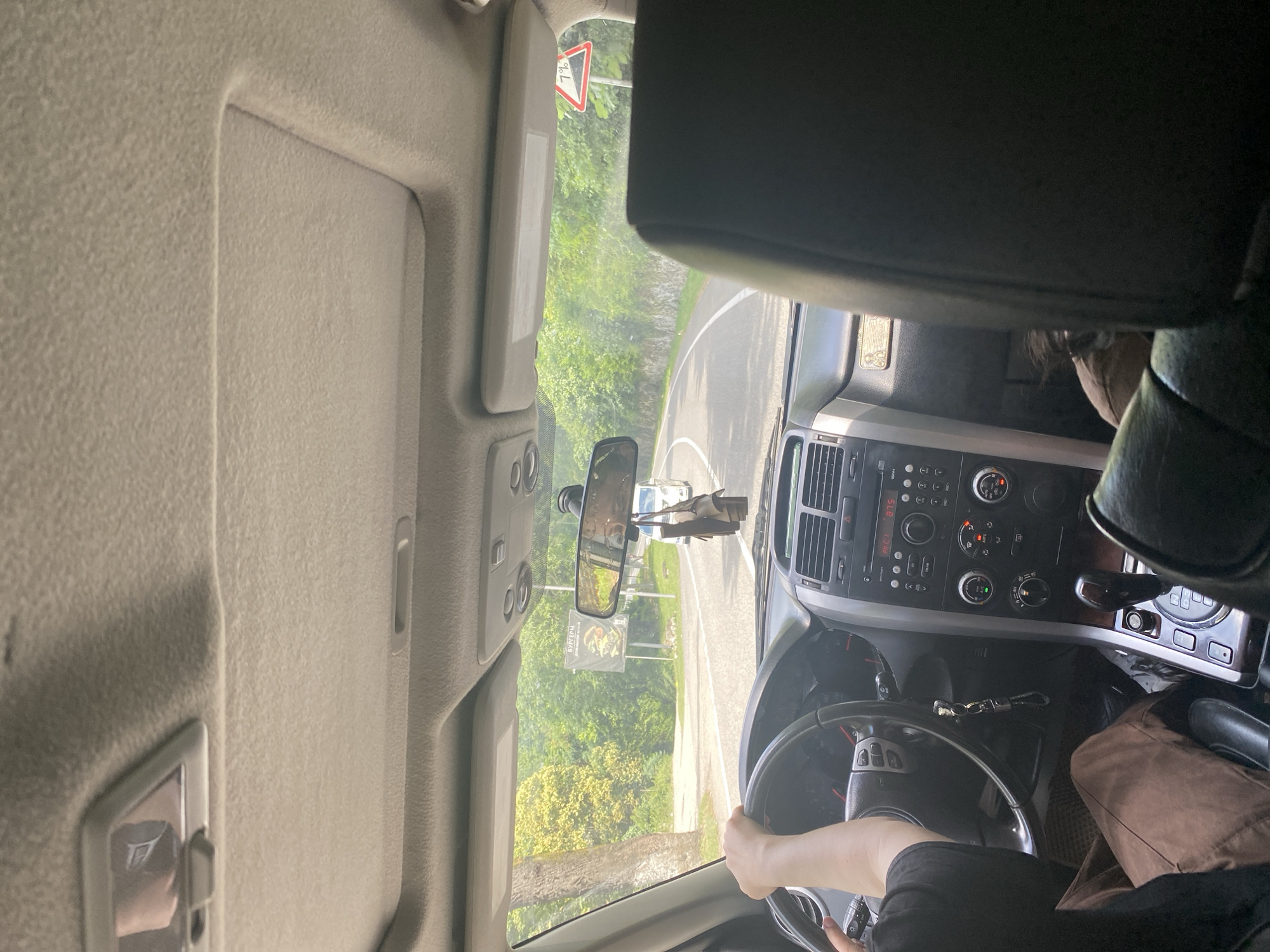 The image shows the interior of a car from the perspective of a passenger, looking forward towards the driver and the road ahead. The driver's hands are on the steering wheel, and they are wearing a dark t-shirt. The car's dashboard is visible, with the radio and climate controls displayed. Outside the car, a road curves through a lush green environment with trees and foliage. There is a billboard visible on the right side of the road with Russian text and an image of a burger. A triangular road sign with "7%" is also present. Two cars are visible on the road ahead, and a third car is partially obscured by the car in front. The scene appears to be set in a rural or semi-rural area, possibly on a sunny day.