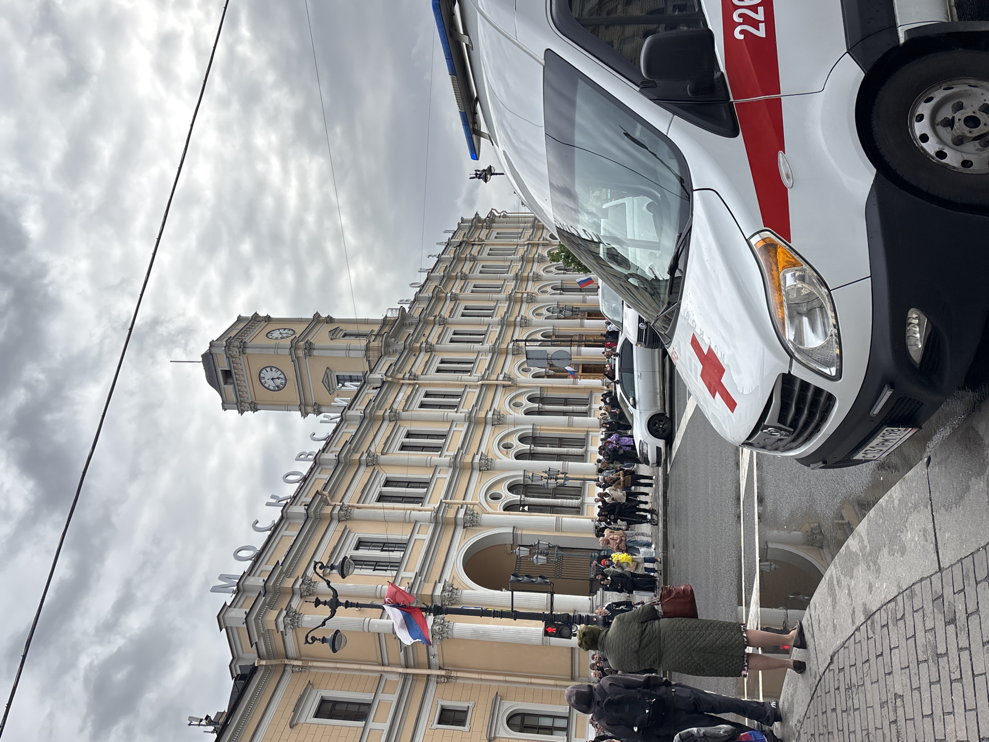 The image shows a street scene in Saint Petersburg, Russia, with the Moskovsky Railway Station in the background. A white ambulance vehicle is parked in the foreground, partially obscuring the view of the station. The station is a large, ornate building with a clock tower and the Russian word "МОСКОВСКИЙ" (Moskovsky) in large letters on the facade. A Russian flag is visible on a lamppost near the station entrance. A line of people is waiting near the station entrance. The sky is overcast. Some pedestrians are on the sidewalk in the foreground. The registration plate of the vehicle in the foreground reads "C331PM198".