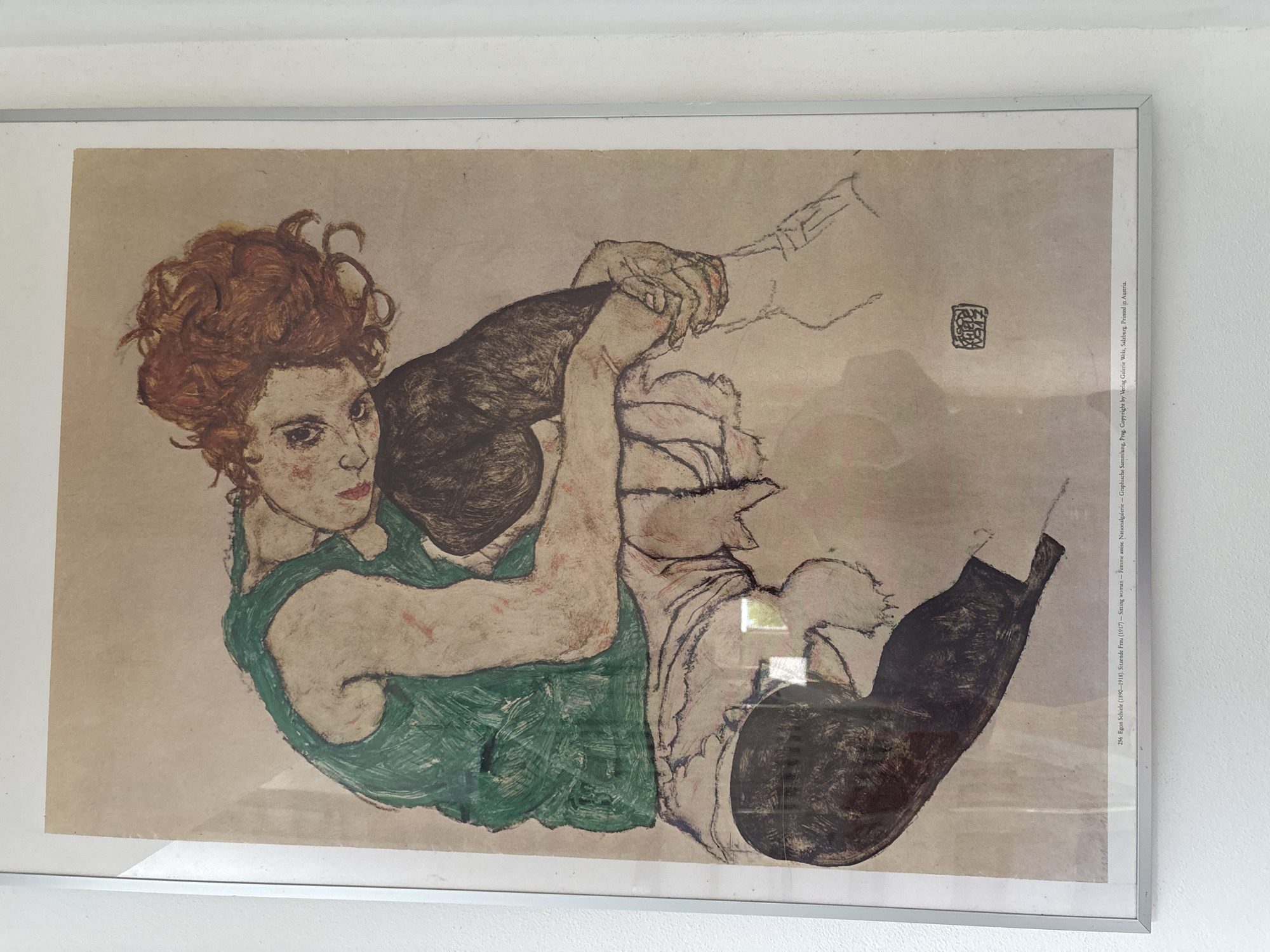 The image shows a framed print of a drawing by Egon Schiele titled "Sitting Woman" (1917). It features a seated woman with reddish hair and a green dress, embracing her legs. The style is expressionistic, with bold lines and distorted proportions. The print includes text identifying the artist, title, location (Nationalgalerie - Graphische Sammlung, Prag), and copyright information. The image was taken in Pisa, Italy, and the location is unknown.