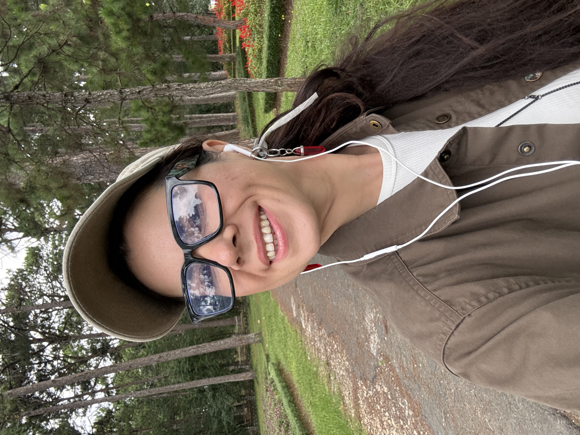 A woman with dark, long hair, wearing a brown jacket, a white ribbed shirt, a tan baseball cap, and black-rimmed glasses, smiles at the camera. She has white earbuds in her ears, one of which has a red pendant earring attached. The woman is outdoors, standing on a path in what appears to be a park. In the background, there are tall pine trees, green grass, and beds of red flowers. This setting is consistent with locations like the Tuyen Lam Lake area or the Da Lat Flower Park in Da Lat, Vietnam, known for its pine forests and cultivated gardens. The path is made of a gravelly, sandy material.