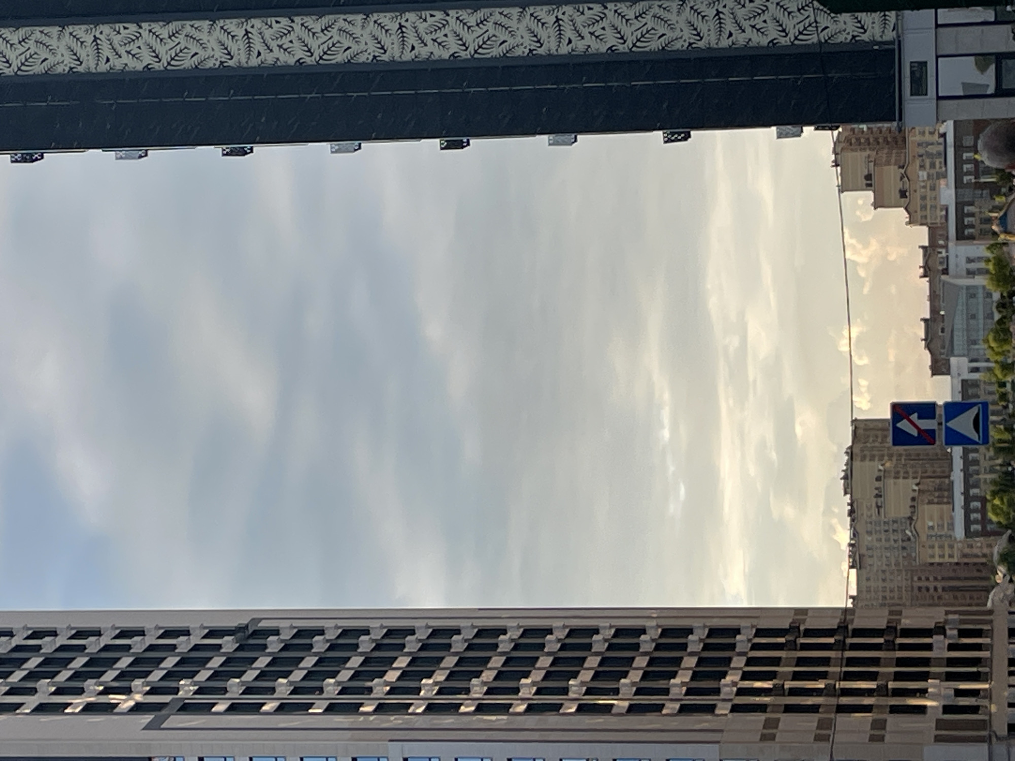 Two modern high-rise buildings frame a view of the sky and other buildings in the background, possibly in Stavropol, Russia. The building on the left is light-colored with a grid pattern of dark windows. The building on the right is dark with a decorative pattern of white leaves or ferns. Between these two buildings, a cluster of lower-rise buildings is visible, some with lighter facades and others appearing older and more textured. The sky is partly cloudy with patches of bright sunlight filtering through. Two traffic signs are visible in the lower middle section of the image. One sign, a blue square with a white arrow pointing upwards and a red circle with a diagonal line, indicates "no entry" or "closed to all vehicles." Below it, another blue sign depicts a triangular shape with wavy lines, likely indicating a speed bump or uneven road surface. The scene appears to be a street-level view, with no prominent people or specific actions captured.