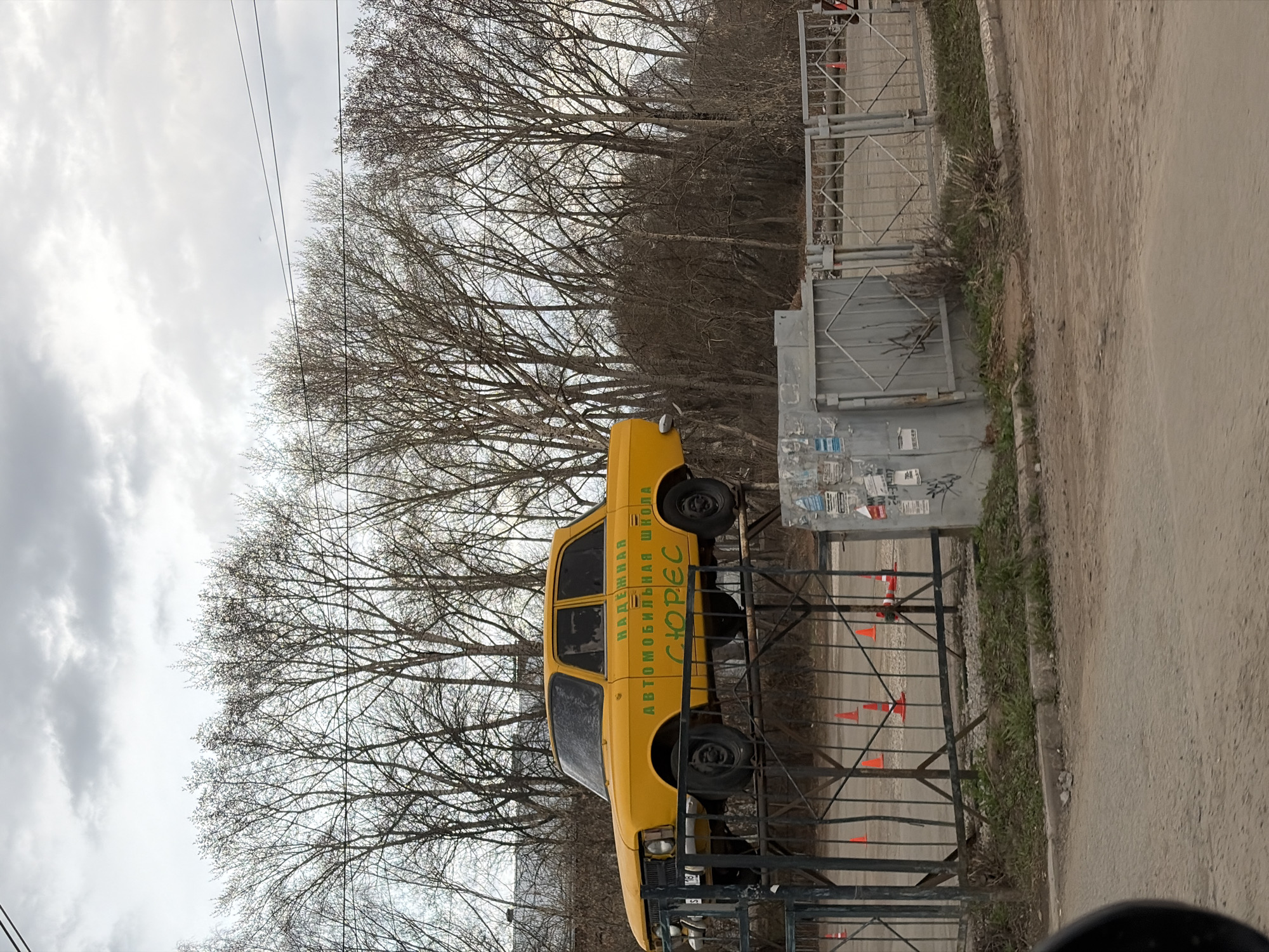 A yellow Moskvich 2140 car is mounted on a metal fence beside a road in Izhevsk, Russia. The car has green writing on its side in Russian, identifying it as a driving school. The fence is made of vertical metal bars. Behind the fence are several orange cones. Behind the car and fence are bare trees without leaves. The sky is overcast. A grey metal structure is to the side of the car. A sidewalk and road are to the right side of the image.