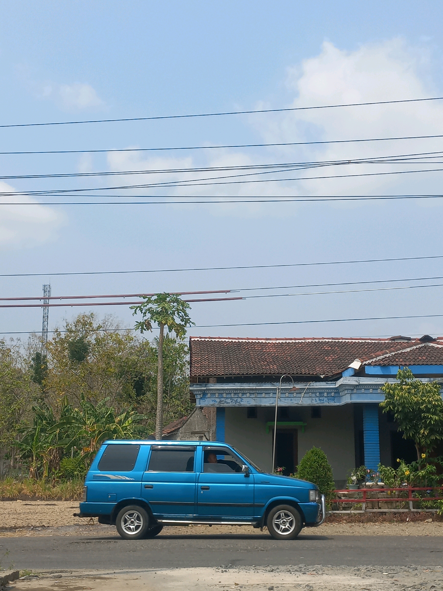 A blue SUV is parked on a dirt road in front of a building with a tiled roof. The building has blue trim and a large open doorway. There are several trees and bushes surrounding the building, and a cell tower can be seen in the distance. The sky is clear and blue with a few white clouds. The scene appears to be in Baturetno, Indonesia, likely during daylight hours.