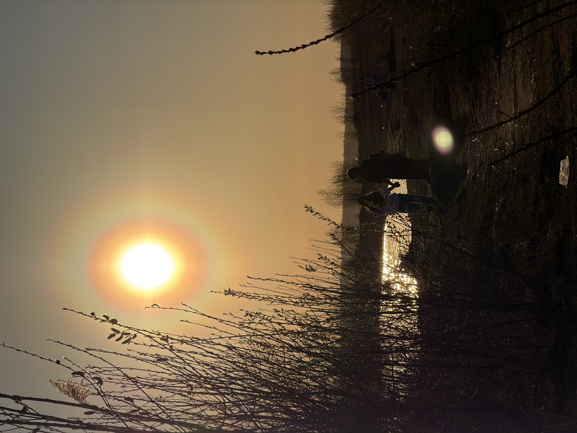 The image shows a waterside landscape at sunset, likely near a body of water in Izhevsk, Russia. The bright sun, surrounded by a halo, is positioned in the upper left. Bare branches of shrubs and trees dominate the foreground. Two people stand near the water's edge on the right side of the frame. The water reflects the sunlight, creating a shimmering effect. The right side of the image features dark land with sparse vegetation, and some litter is visible.