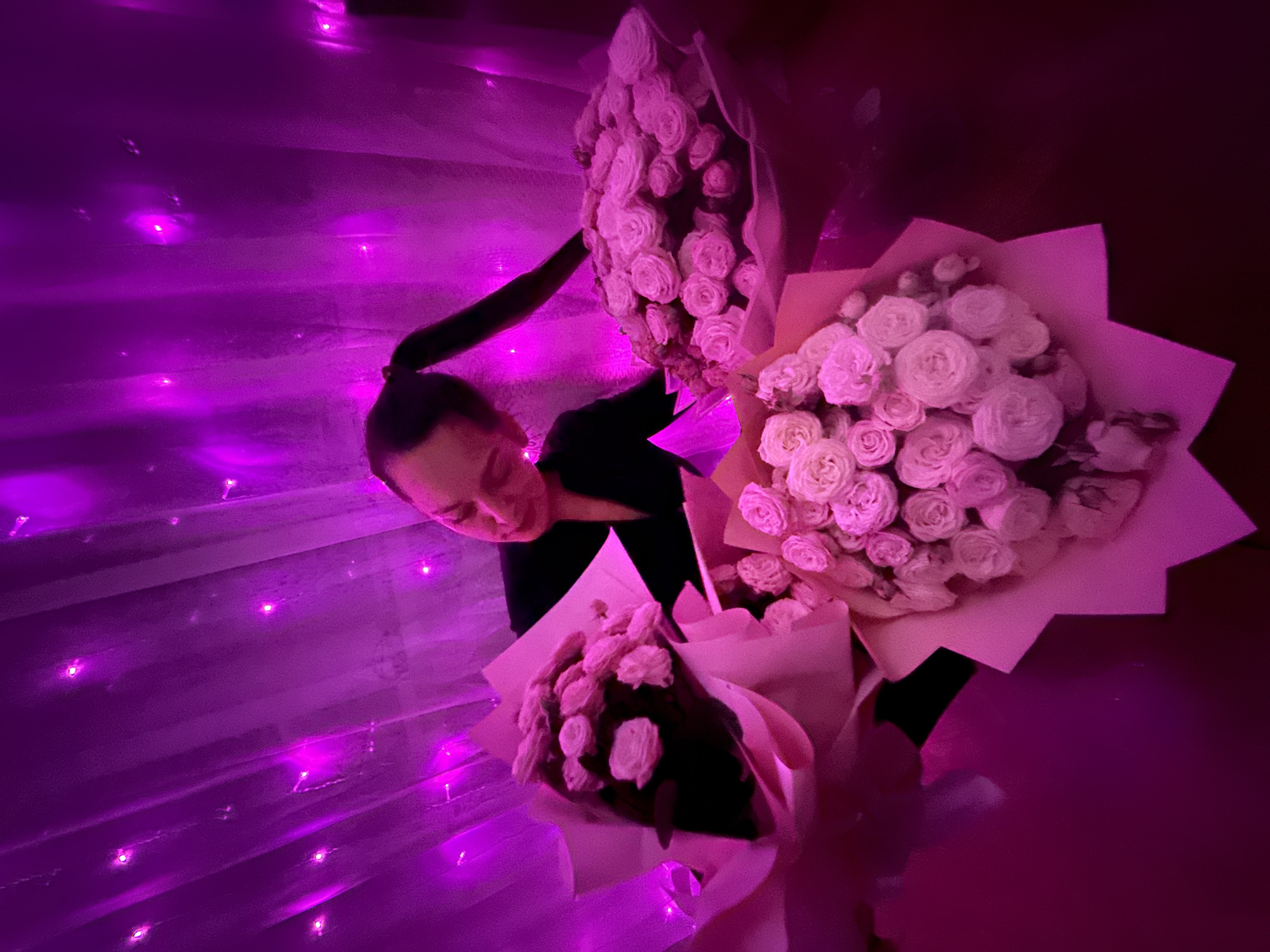 A woman with a ponytail in a black shirt holds three bouquets of roses. The scene is lit with bright purple light, suggestive of a celebratory or decorative setting. The roses are of a light color, wrapped in paper. In the background, there's a wall or backdrop with what appears to be lights. The exact location in Chaikovsky, Russia, cannot be determined from the image.