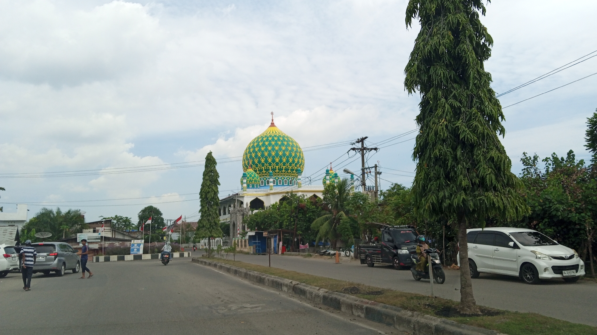 The image shows a street scene in Lhokseumawe, Indonesia. A mosque with a distinctive green and gold dome is visible in the background, surrounded by trees. Several vehicles are on the road, including cars and motorcycles. People are walking and riding on motorcycles. The sky is cloudy. The overall impression is of a typical day in a Southeast Asian city.