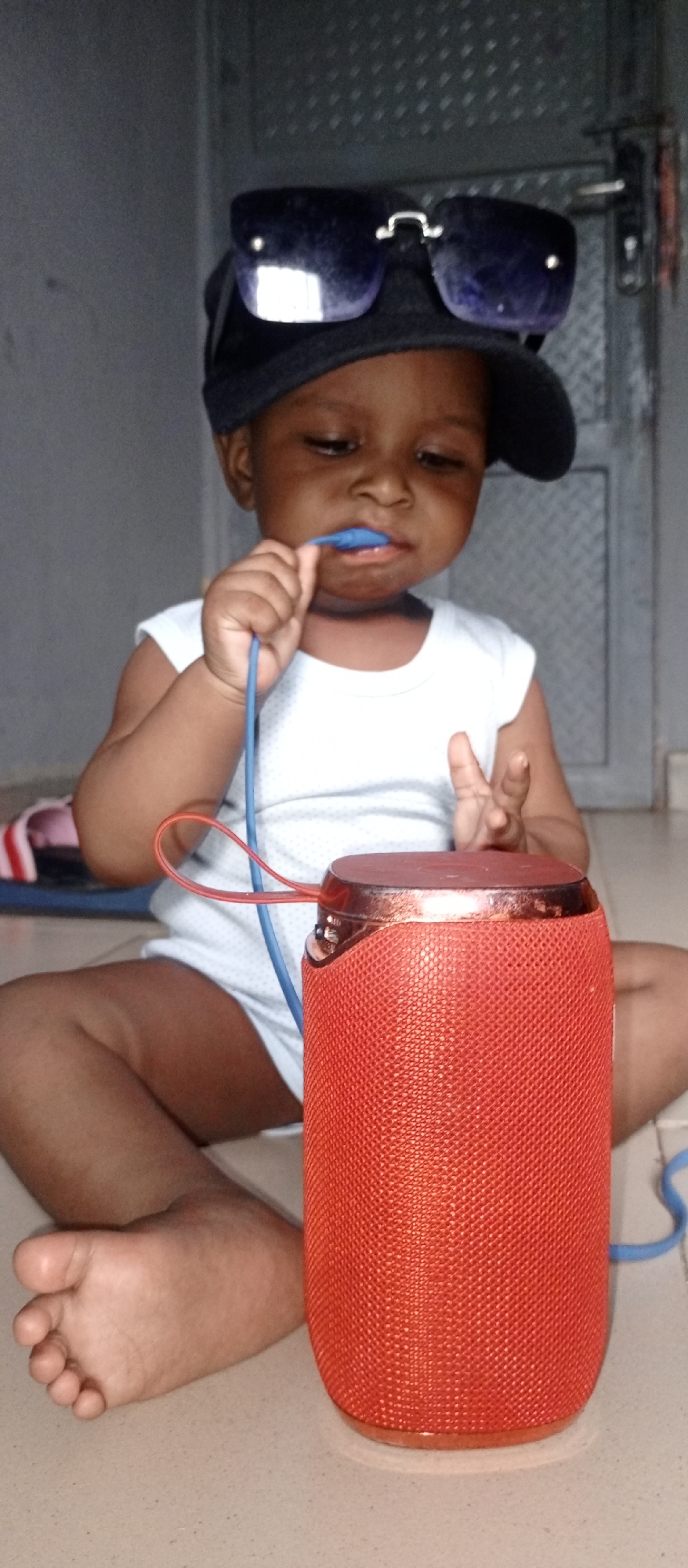 A toddler sits on a light-colored tiled floor, next to a cylindrical red speaker. The child is wearing a white onesie, a dark baseball cap, and sunglasses perched on top of the cap. The toddler is holding a blue cord in their mouth and gesturing with their right hand towards the speaker. A red sandal lies on the floor behind the child. A gray wall and a door are visible in the background. The location is in the city of Unknown, Unknown.