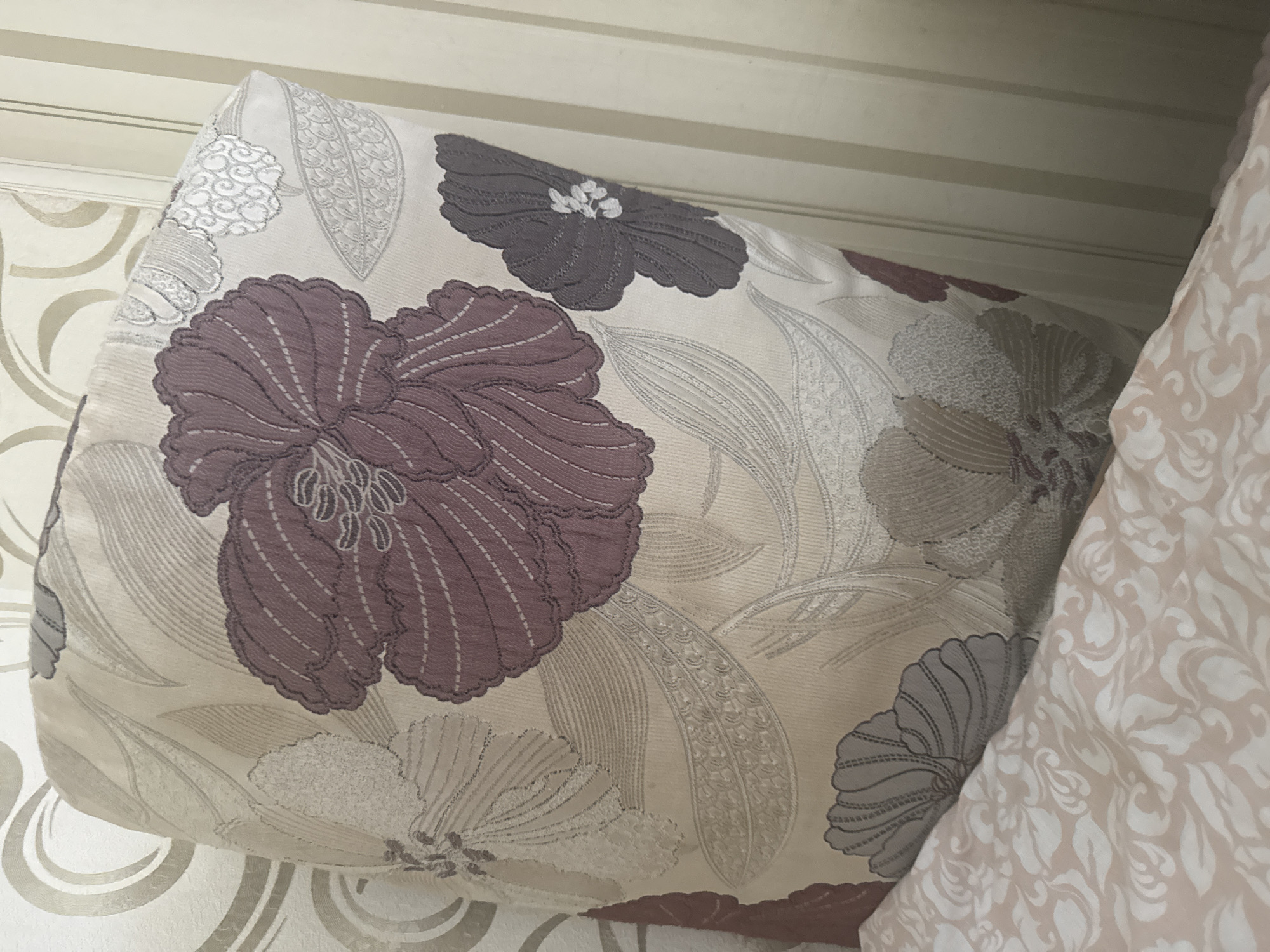 The image shows a close-up of a pillow with a floral pattern, placed on a bed. The pillow has large, stylized flowers in shades of mauve, grey, and off-white, with intricate detailing. The fabric of the pillow appears to be textured. The pillow is leaning against a wall with wallpaper that has a subtle swirling pattern. To the right of the pillow, a vertical striped wallpaper or wall covering is visible. In the foreground, a portion of the bed is visible, covered with a pink and white patterned fabric that also features a swirling floral motif. The lighting in the image is soft, suggesting an indoor setting. There are no people or animals visible in the frame, and no actions or interactions are depicted. The image does not contain any identifiable landmarks or features that would allow for its location to be specified as Poltava, Ukraine.