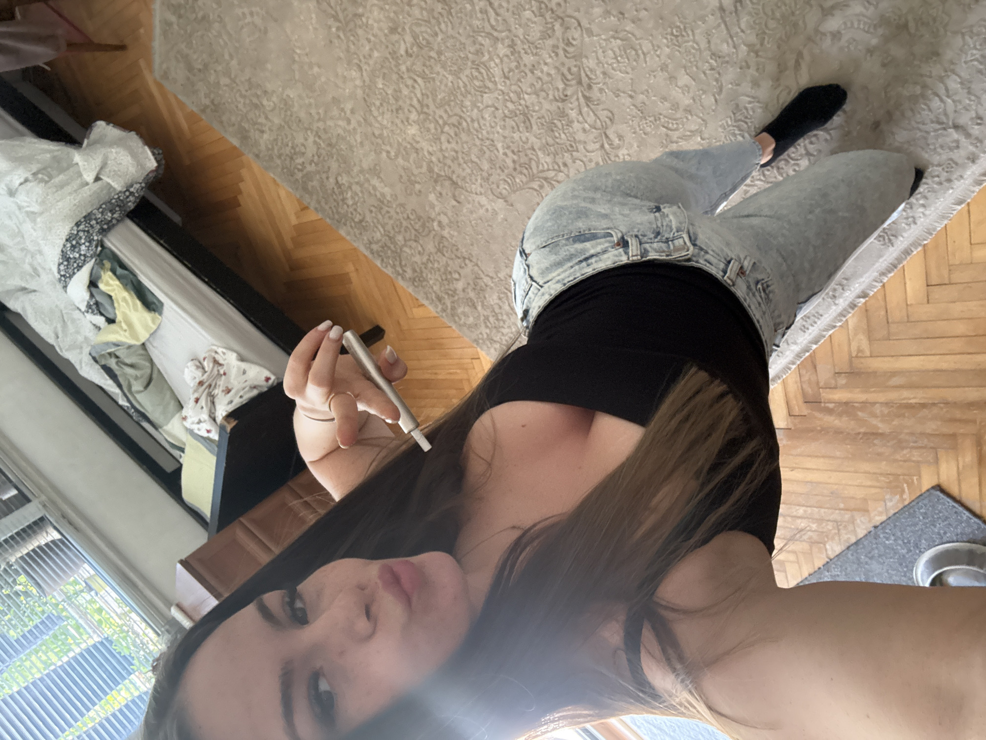 A female is seen in a selfie. She is wearing jeans, a black shirt, and has brown hair. She is holding a small silver object in her right hand. The background includes a bed with linens, a rug on a herringbone wood floor, and a window. There is no indication of location in Plovdiv, Bulgaria.