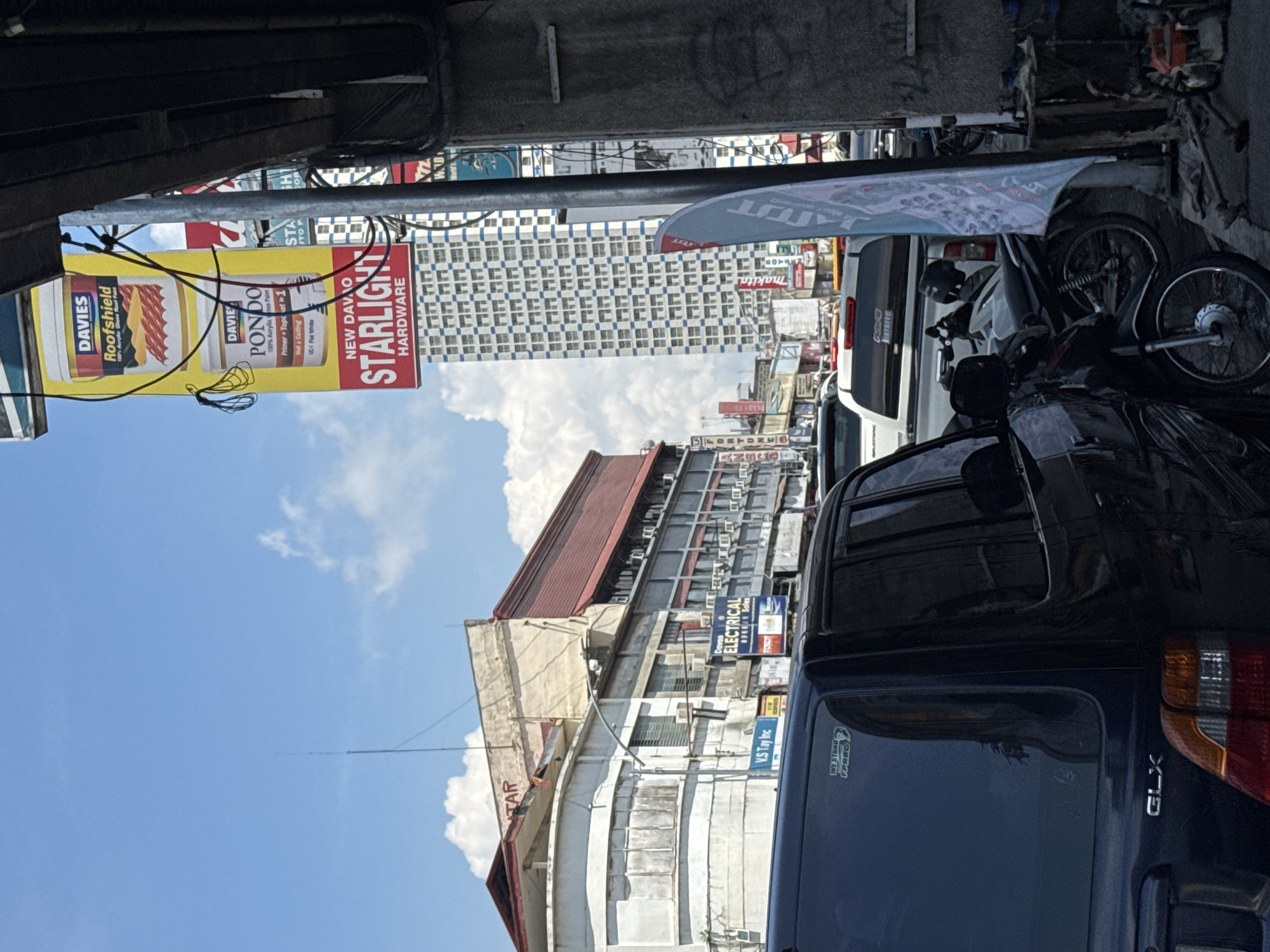 A street scene in Davao City, Philippines, features a variety of commercial establishments and vehicles under a bright, cloudy sky. A tall, modern building with a blue and white facade stands prominently in the background, contrasting with older, multi-story structures to its left that house businesses like "DAVIES Roofshield," "PONDO," and "STARLIGHT HARDWARE." Signs for "Davao ELECTRICAL" and "V.S Tay Inc." are also visible.
In the foreground, several vehicles are parked along the street. A black SUV with the "GLX" model designation is parked on the left, partially obscuring a white pickup truck behind it. Multiple motorcycles are also present, some parked and others appearing to be in use. The street appears to be a bustling commercial area with a mixture of modern and older architecture. The overall impression is that of a typical urban street in the Philippines.