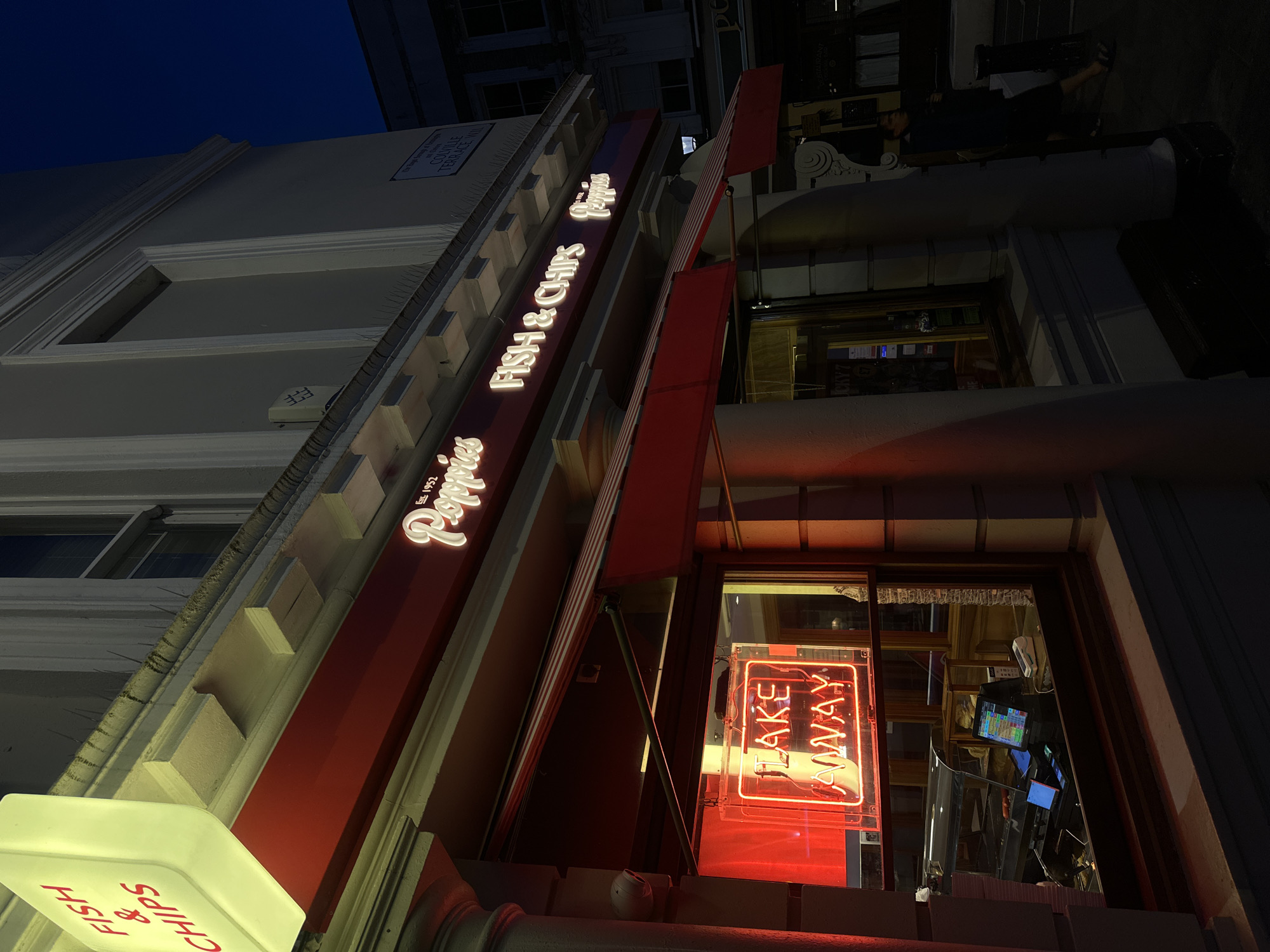 The image depicts the exterior of "Poppies Fish & Chips" in London, United Kingdom, at night. The restaurant's sign is illuminated. A red neon "TAKE AWAY" sign is visible inside the window. The architecture of the building is visible.