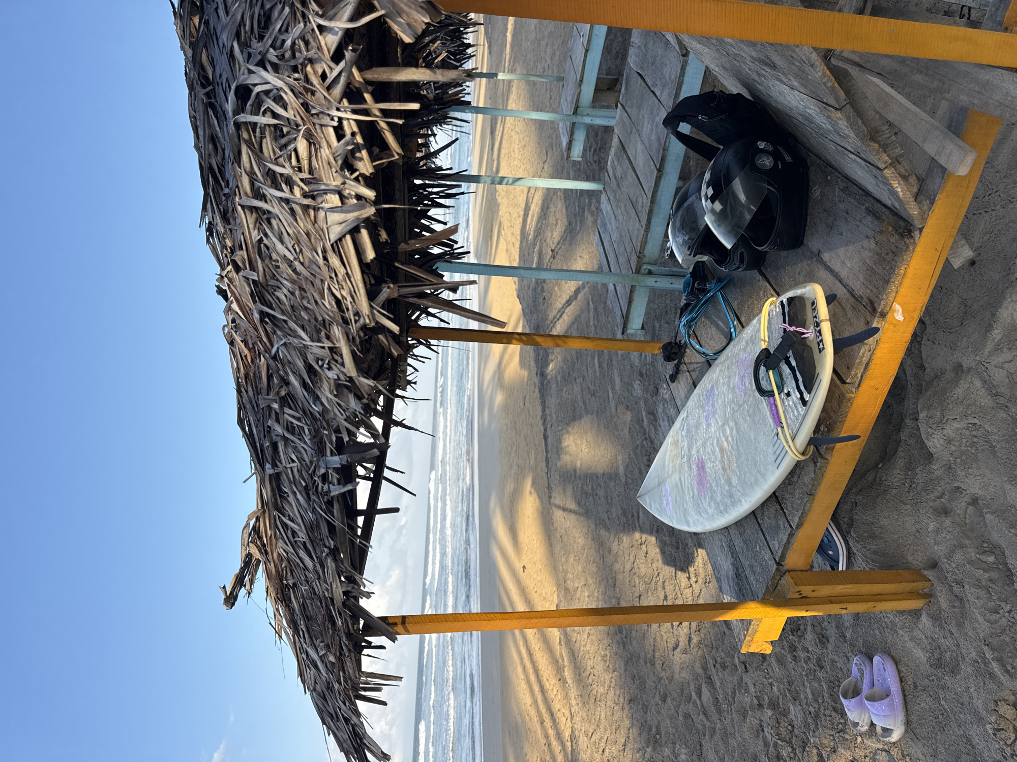 A surfboard, two helmets, and a pair of sandals are arranged on a wooden platform under a thatched-roof shelter on a beach in Kathaluwa, Sri Lanka. The surfboard is light-colored with purple markings and is positioned horizontally on the platform. Two black helmets, one with its visor up, sit next to the surfboard. A pair of light purple sandals rests on the sand to the left of the platform. The shelter, made of dried palm fronds, provides shade, casting long shadows on the sandy beach. In the background, the ocean stretches to the horizon under a clear blue sky with a few wispy clouds. The overall scene suggests a moment of rest or preparation for water sports.