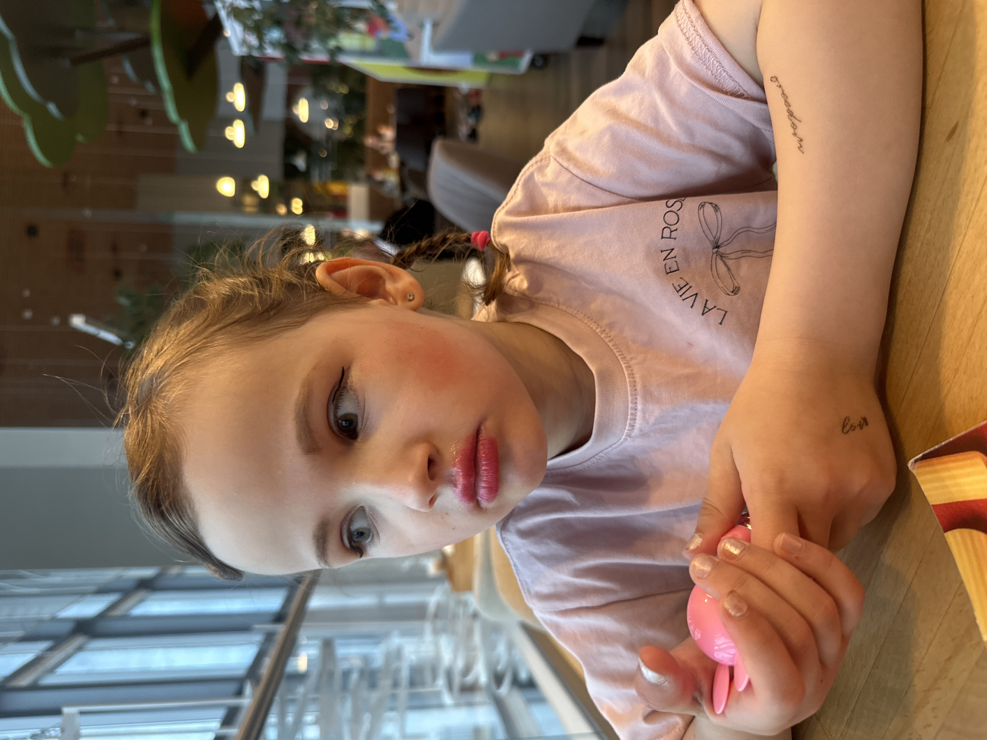 A young female child is depicted indoors, seated at a light-colored wooden table in what appears to be a cafe or restaurant in Khabarovsk, Russia. The child, shown in a frontal-left profile, has light-colored hair pulled back, with a small braid secured by a pink elastic visible on the right side. Her left earlobe contains a small stud earring. She exhibits applied makeup, including prominent black winged eyeliner, pink-red blush on her cheeks, and glossy bright pink lipstick on her pursed lips. Her gaze is directed towards the left, looking out through large glass windows that reveal a blurred urban or street scene. She wears a light pink short-sleeved t-shirt with the text "LA VIE EN ROSES" and a bow graphic printed on it. Her fingernails are painted with a light, shimmery polish. She holds a small, bright pink, plastic object with two ear-like protrusions in her hands. A temporary tattoo-like script reading "love" (inverted) is visible on the back of her right hand, and another script reading "freedom" (inverted) is on her left forearm. In the background to the right, the interior of the establishment shows warm yellow light fixtures, green tree-like decorative elements suspended from the ceiling, and indistinct shapes of other patrons and furniture. A portion of a yellow and red striped package is visible on the table in the bottom right corner of the frame.
