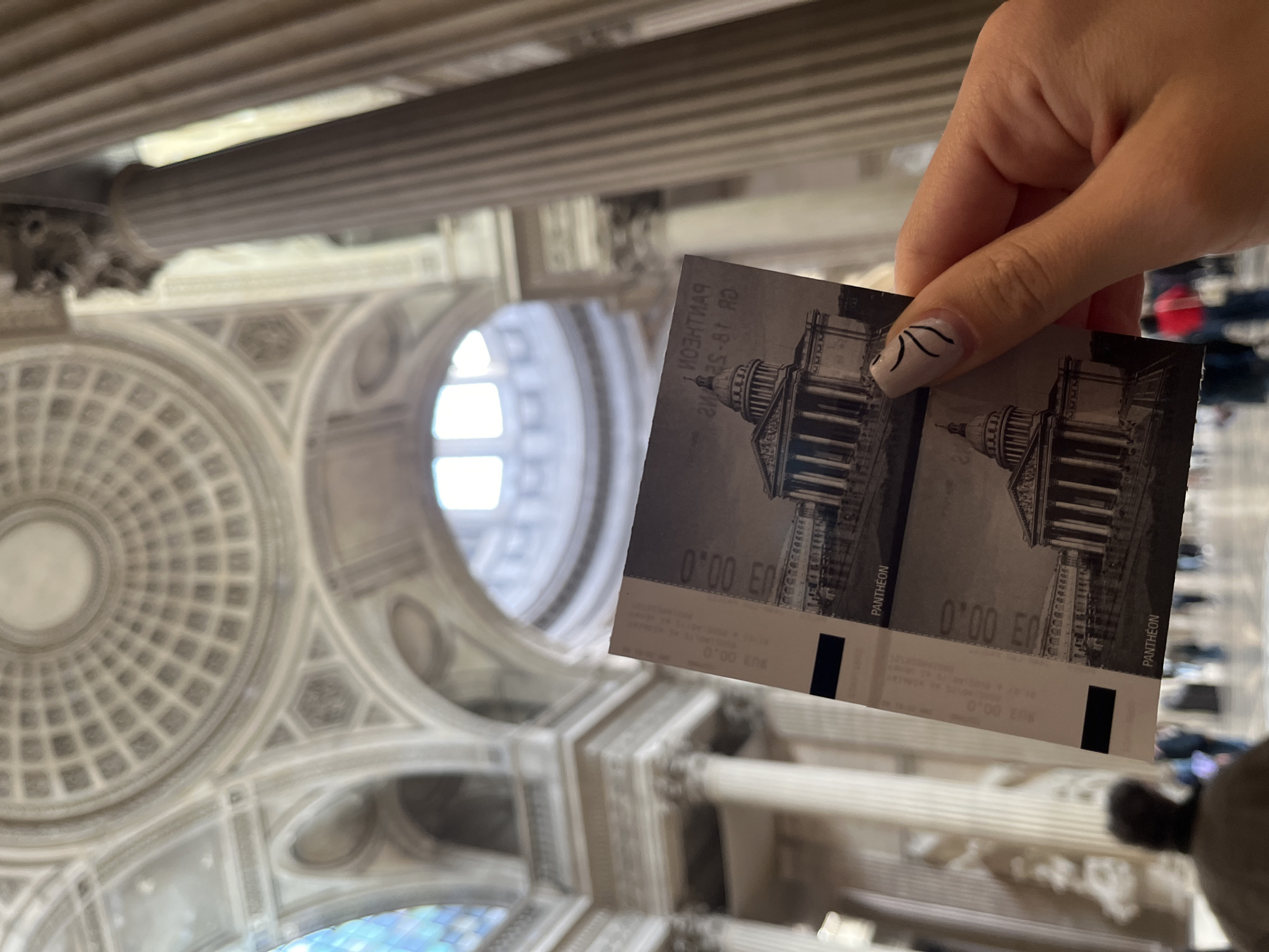 A close-up photograph features a person's hand holding two entrance tickets in the foreground, set against the opulent interior of the Pantheon in Paris, France. The hand, with fair skin and a fingernail adorned with two black wavy lines on a light base, presents the tickets directly to the viewer.
Each gray-toned ticket displays an illustration of the Pantheon's classical facade with its iconic columns and dome. Visible text on the tickets includes "PANTHEON" and the price "0'00 EU," implying complimentary admission. Smaller, partially obscured text such as "24.20.01 RD" is also discernible, likely indicating a date or ticket identification.
The background reveals the grandeur of the Pantheon's architecture, captured from an upward-looking perspective. A large, ornate, coffered dome with intricate geometric patterns fills a significant portion of the scene. Fluted columns and detailed arched structures, typical of neoclassical design, frame the space. Bright natural light streams through arched windows in the mid-ground, suggesting the image was taken during the daytime. The overall scene depicts a visitor documenting their experience inside this historic Parisian monument.