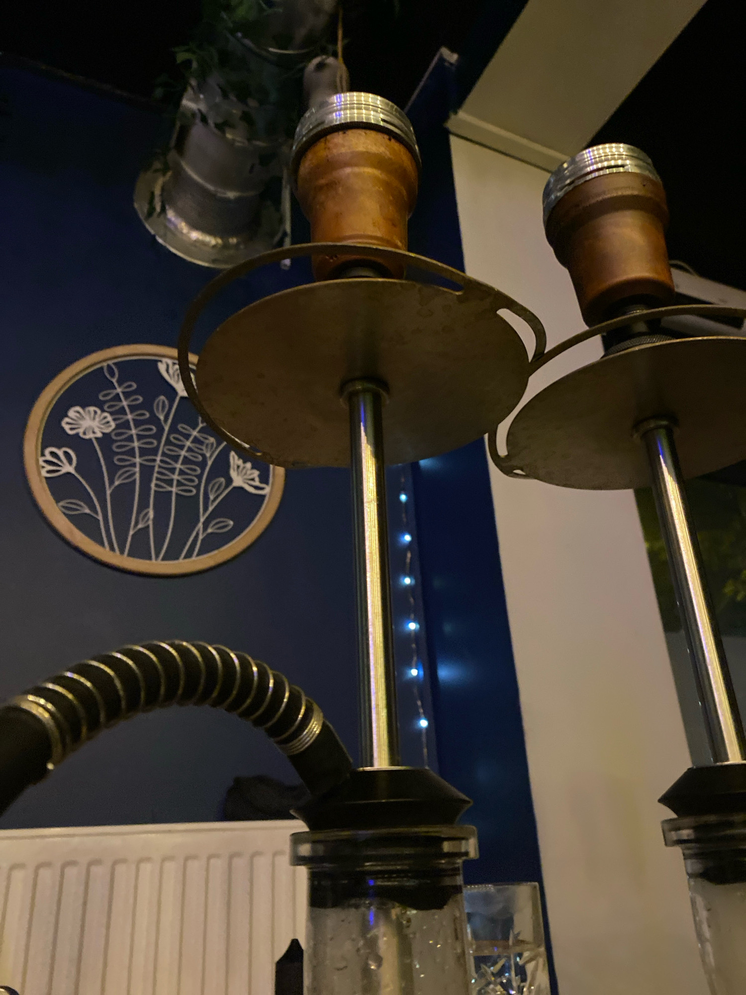 Two hookah pipes are visible in the foreground, one closer than the other, positioned in what appears to be an indoor establishment. The pipes are made of metal and wood, with ornate details on the bowls and stems. A black, ribbed hose extends from the left hookah. In the background, a circular wall decoration with a floral motif is visible against a dark blue wall. A metal radiator is present in the lower left of the frame, suggesting a heated indoor environment. String lights with small blue bulbs are draped along the wall. The overall ambiance suggests a lounge or bar setting. The location is identified as Gliwice, Poland.