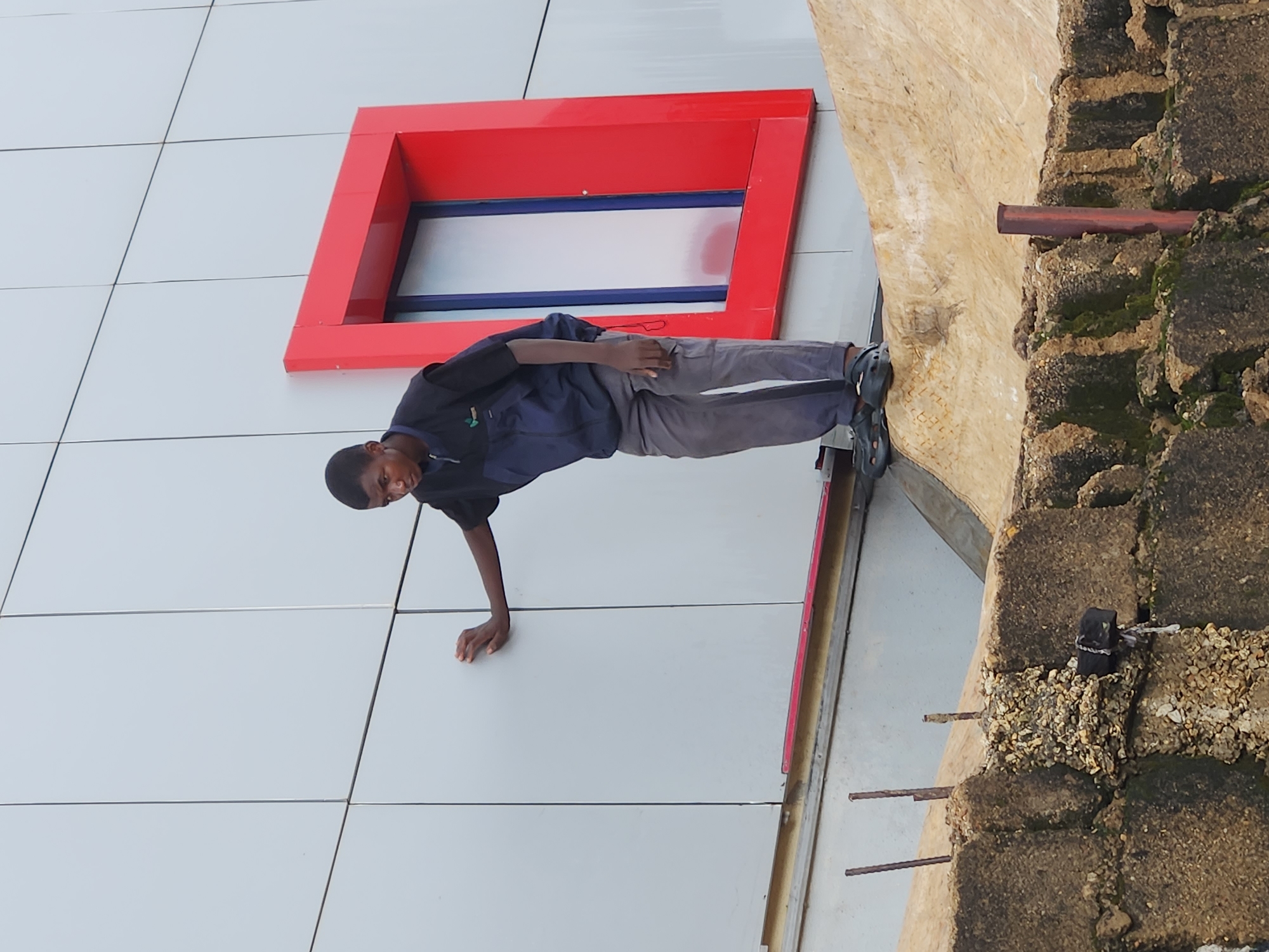 A young Black male is captured in a dynamic pose, leaning against a modern building with a distinctive red-framed window. He is dressed in a dark blue jacket and grey pants, with black athletic shoes on his feet. His right hand is braced against the wall for support, while his left hand rests on his hip. The building features a white tiled facade, creating a clean and geometric backdrop. To the right of the building, a more rustic, textured wall of concrete blocks and gravel is visible, with moss growing on some surfaces. A rusty metal pipe juts out from this older wall. The lighting suggests it could be daytime, with no strong shadows indicating a specific time. The overall scene has an urban or industrial feel, but the specific location is not identifiable. The person's expression is neutral, and the image does not convey any overt emotion. There is no visible text in the image.