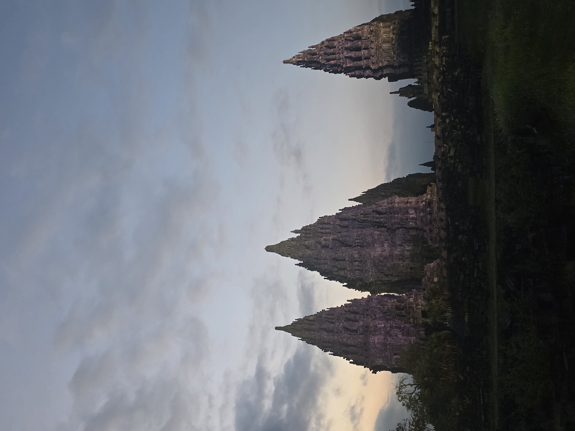 The image captures the Prambanan Temple complex in Bokoharjo, Indonesia, against a backdrop of a cloudy sky at dusk or dawn. Three main temples dominate the foreground, their stone structures and tiered roofs clearly visible. The lighting casts a soft glow on the temples, highlighting their intricate carvings. To the right, vegetation and some portions of other temple structures are visible.