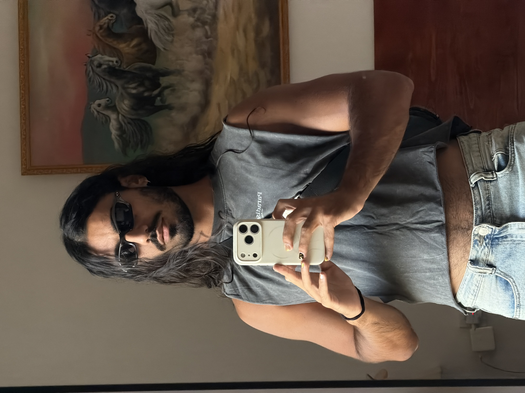 A person with long dark hair and a beard, wearing sunglasses, a grey sleeveless crop top, and light blue jeans, stands in front of a mirror, taking a selfie with a white iPhone. Behind them, a painting of galloping horses hangs on the wall. The setting appears to be an indoor space, possibly a bedroom or dressing area, with a white power outlet visible on the wall. There is no discernible indication of the specific location in North Kuta, Indonesia.