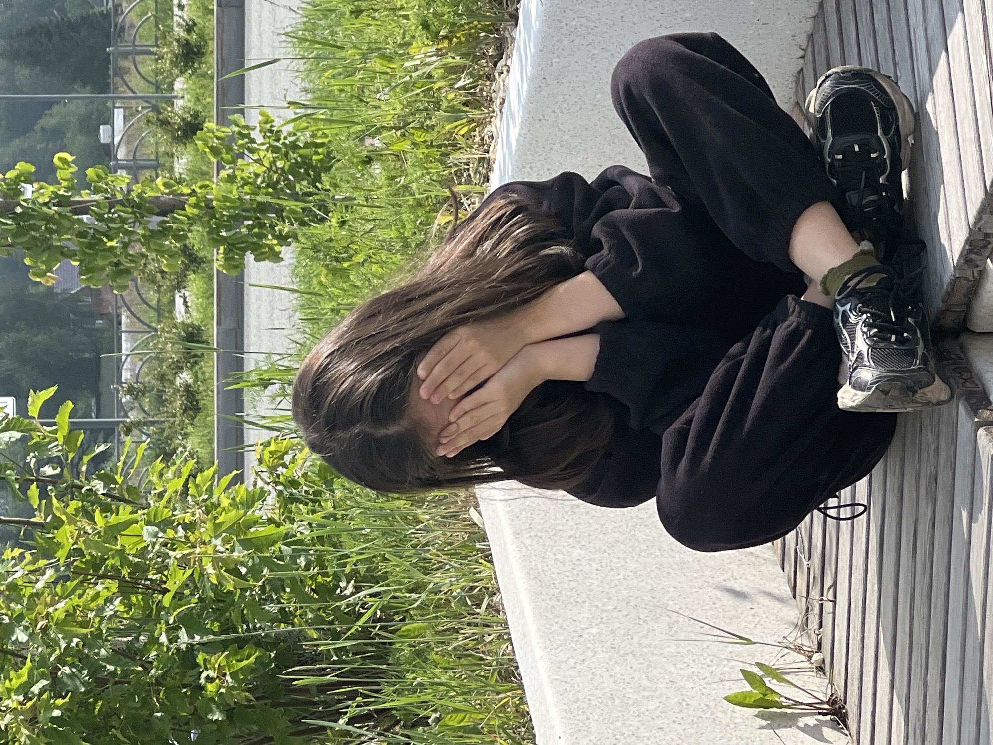 A young individual with long dark brown hair is seated cross-legged on a modern outdoor bench in an urban green space. The person is dressed in a dark, likely black, long-sleeved top and matching sweatpants, paired with dark athletic sneakers featuring white soles and green ankle socks. Their hands are covering their face. The bench consists of wooden slats for the seating surface and a light-colored, possibly concrete or composite, backrest/side with a granular texture.
Behind the bench, a strip of unkempt green grass and various small plants is visible, including a small broad-leafed plant growing from a crevice in the bench structure. Further back, two trees or large bushes are present: a dense, leafy green bush on the left and a more slender, sparsely-leafed tree on the right. Beyond the greenery, a paved area, likely a road or pedestrian path, can be seen. In the far background, a dark metal fence with repeating arched sections runs horizontally, separating the foreground from more distant, indistinct greenery and structures, possibly residential or public buildings. The scene is illuminated by strong sunlight, casting visible shadows and suggesting a clear daytime environment. The location is an unspecified public green area within the city of Yekaterinburg, Russia.