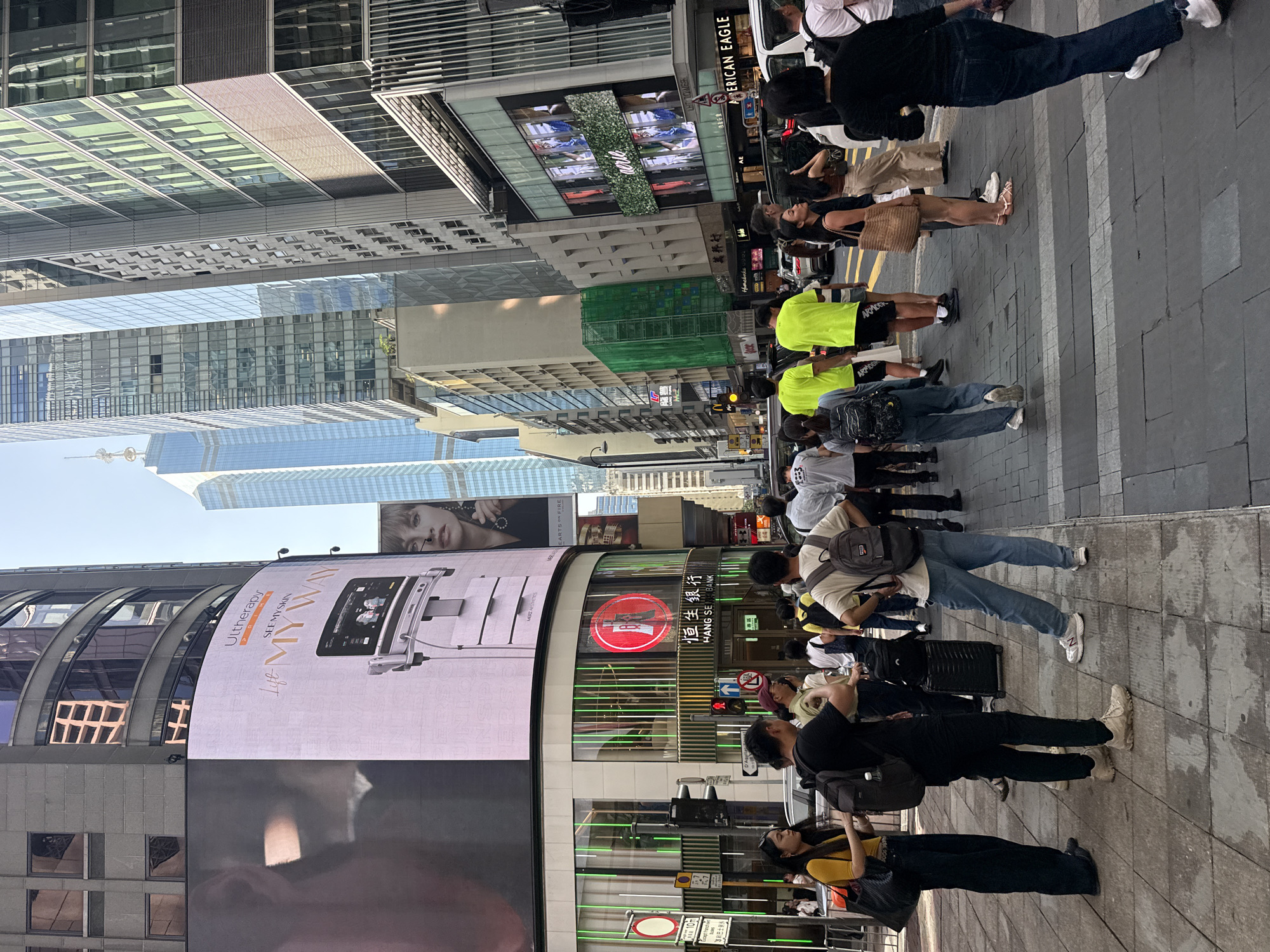 This is a street-level view of a busy urban environment in Hong Kong, China, likely during daytime. The perspective is from a pedestrian perspective, looking down a street lined with tall buildings.
In the foreground, several pedestrians are visible. On the left, a woman wearing a yellow top and dark pants is standing, holding a large black bag. Next to her, a man in a black t-shirt and dark pants is also standing, carrying a black backpack. Further back and to the right, a group of people are walking or standing on the sidewalk, some wearing bright neon green shirts. One man in the center foreground is wearing a light-colored t-shirt, blue jeans, and a backpack, and is standing with his hands on his hips. To his right, a woman in a brown top and beige pants is holding a woven bag.
The street features a pedestrian crossing with painted white stripes. Several vehicles, including cars and a white van, are present on the road. Traffic lights are visible, indicating the presence of intersections.
The buildings flanking the street are characteristic of Hong Kong's dense urban landscape. A large digital billboard is prominently displayed on the side of a building on the left, showing an advertisement for Ultherapy. Below the billboard, a storefront with green lighting and a red circular logo is visible, with signage in Chinese characters and the words "HANG SENG BANK." On the right side of the street, other building facades are visible, some with glass windows reflecting the sky and surrounding structures, and an "AMERICAN EAGLE" storefront is identifiable. Another large digital screen displays imagery of people. The sky is clear and blue.