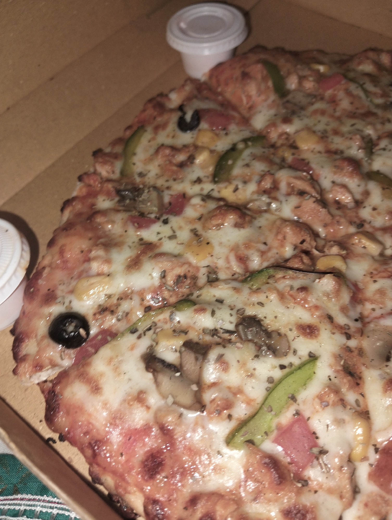 A close-up shot of a pizza in a cardboard box, with two small white containers placed near the top-left corner. The pizza has visible toppings including cheese, mushrooms, black olives, green peppers, diced red peppers, and what appears to be shredded chicken or a similar meat product. Some herbs or spices are sprinkled across the pizza. The scene likely represents takeout or delivery food. There is no indication of location, and therefore the image cannot be said to have been taken in Dhaka, Bangladesh, or any specific place.