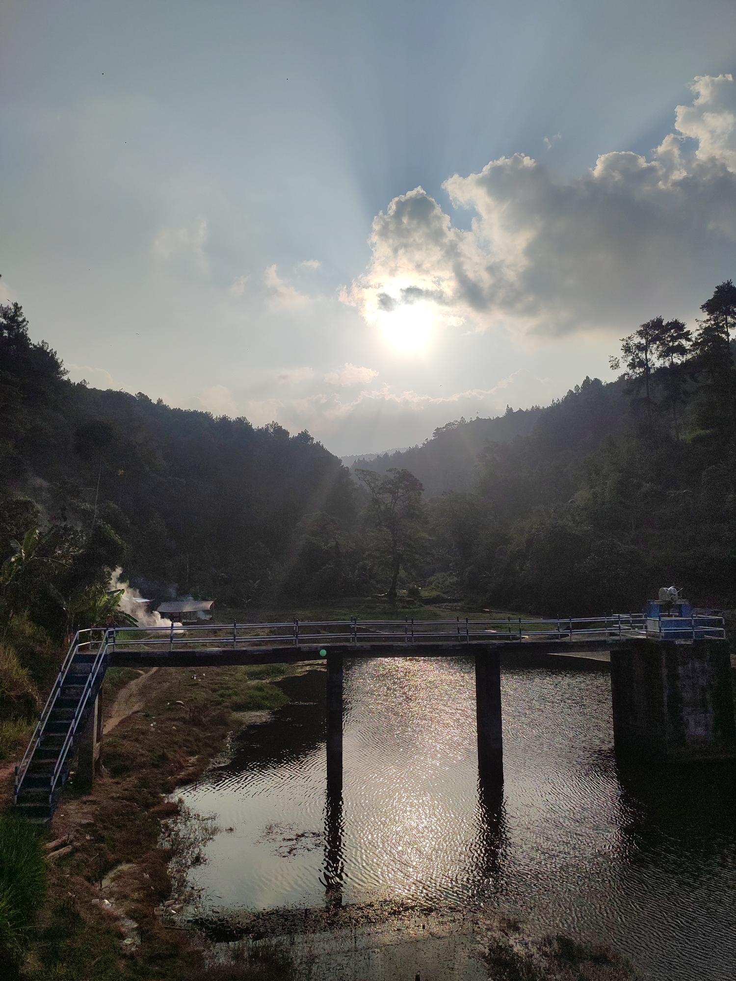 The image depicts a scenic view of a bridge crossing a body of water, surrounded by lush green hills and trees. The sun is setting, casting a warm golden glow across the scene, with rays of light piercing through the clouds. On the left side of the bridge, a small structure with smoke rising from it is visible, suggesting human activity. The water reflects the sunlight and the surrounding landscape, creating a shimmering effect. The bridge itself appears to be made of concrete and has railings on both sides. The overall atmosphere is serene and tranquil. The image is captured at a slight angle, looking across the bridge towards the hills. It is not possible to determine the exact location within the city of Joho, Indonesia, from the image alone.