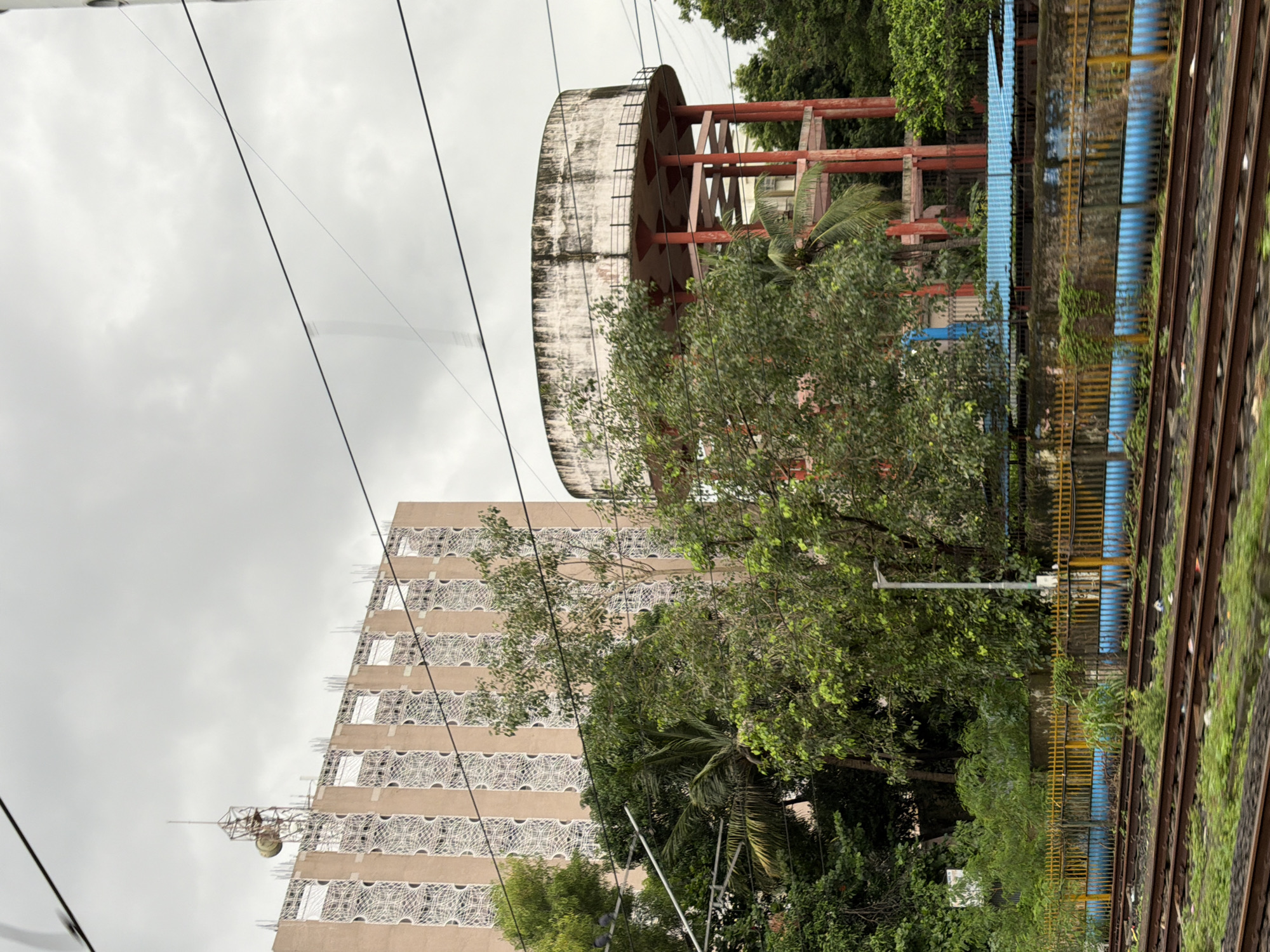The image captures a view from a train traveling through Mira-Bhayander, India. In the background, a tall, multi-story building with a light brown facade and decorative white patterns on its vertical sections stands against a cloudy sky. A prominent telecommunications tower with an antenna is situated on top of the building. To the right of the building, a large, circular water tank is elevated on red supporting pillars. Lush green trees and foliage are abundant in the foreground and middle ground, obscuring parts of the structures. Railway tracks dominate the lower portion of the image, with a yellow fence and blue pipes visible behind them. The overall scene depicts an urban landscape with a mix of infrastructure, natural elements, and the signs of active train travel.