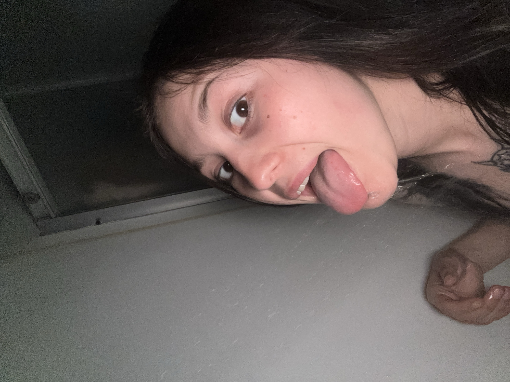 The image depicts a young woman in what appears to be a bathroom setting. The woman is sticking her tongue out and looking towards the camera. She has dark hair and light skin. A portion of her upper body is visible, revealing a tattoo on her shoulder. Her hand is also visible at the bottom right of the image. The background includes a white wall or surface, possibly part of a bathtub or shower enclosure. A dark area, possibly a window or opening, is visible at the upper left of the image. There is no indication of the location being in the city of Фоки, Russia.