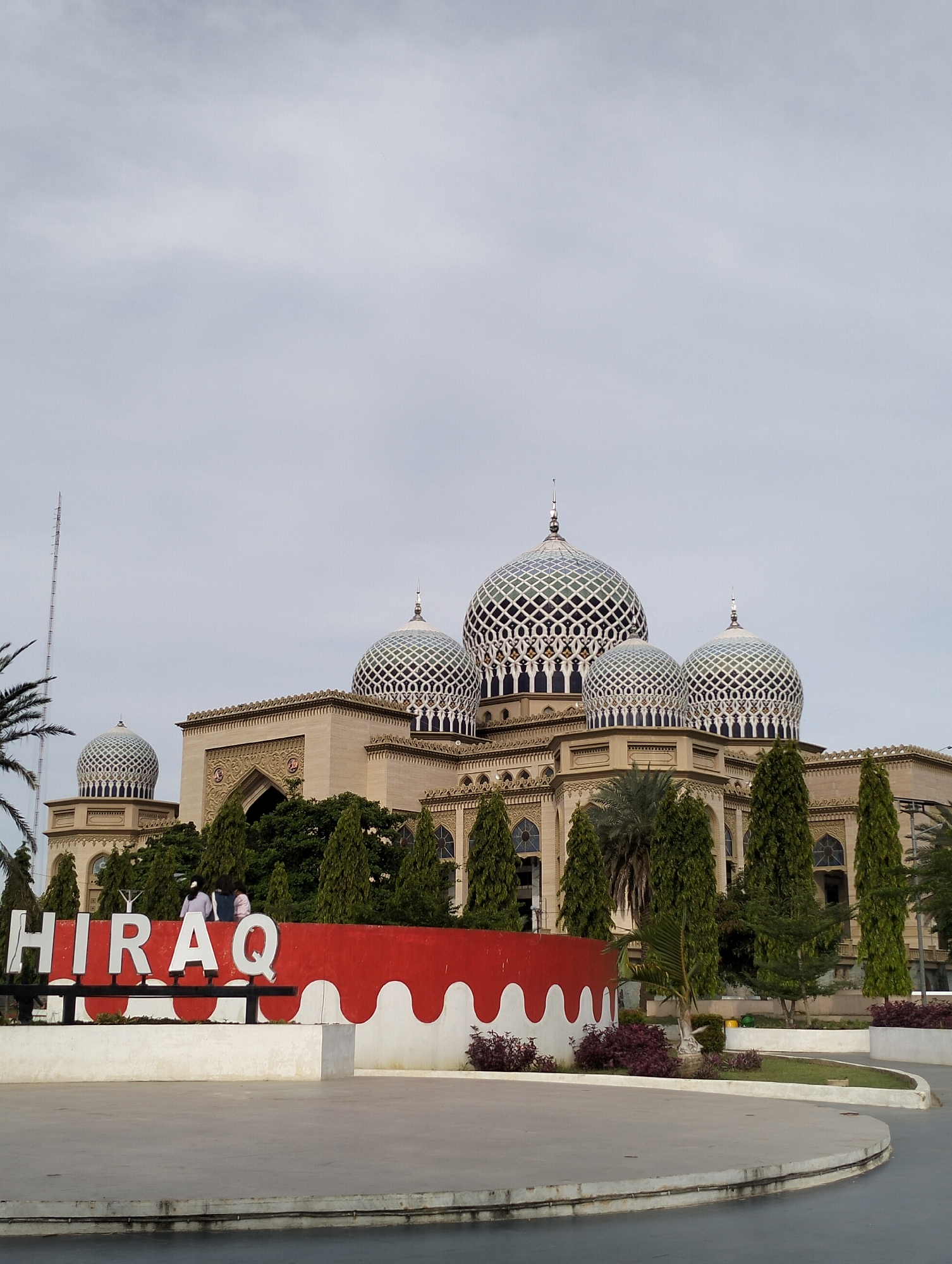 The image depicts a view of the Islamic Center mosque in Lhokseumawe, Indonesia, against a cloudy sky. In the foreground, a large sign spells out "HIRAQ" in block letters, with a red and white decorative base. Two figures, possibly people, are situated on the sign. The mosque, featuring multiple domes with geometric latticework patterns, rises behind the sign, partially obscured by trees and other greenery. A tall antenna is visible on the left side. The foreground also includes a paved area.