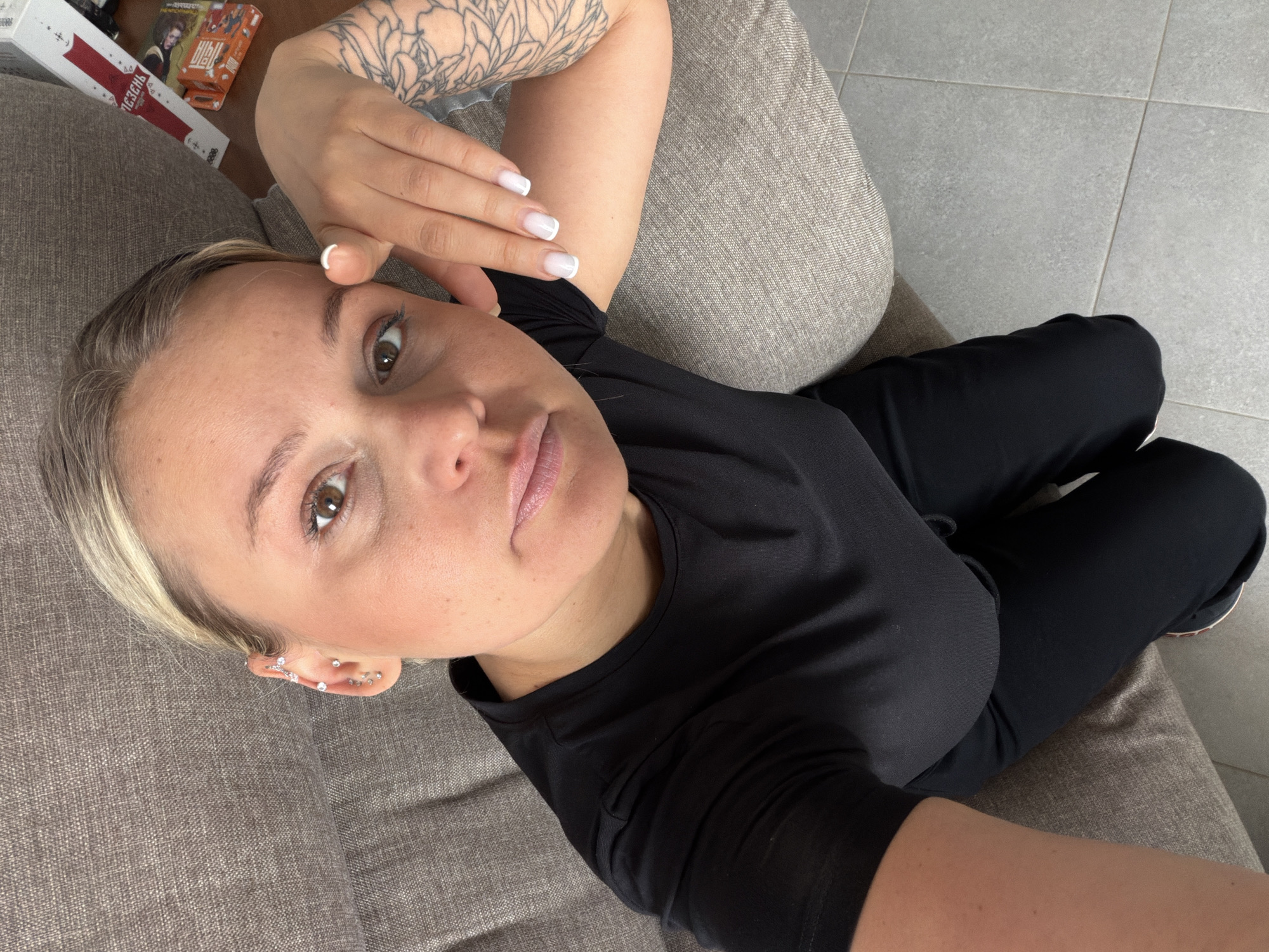 A fair-skinned woman with short blonde hair and multiple ear piercings is reclining on a grey fabric couch. She is wearing a black T-shirt and black pants. Her left arm, which has a tattoo, is raised with her hand near her forehead. Her nails are painted white with a nude base. Several objects are visible near the top left corner of the image. The location appears to be indoors on a tiled floor, but the specific city of Sarapul, Russia, cannot be determined from the image.