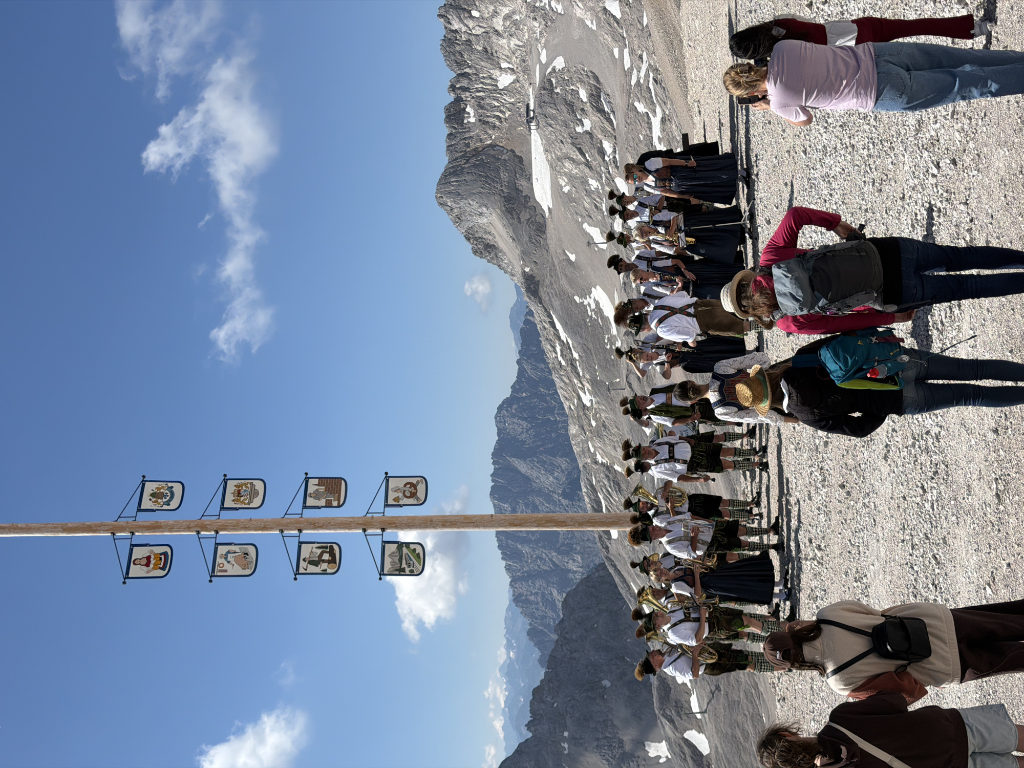 In Leutasch, Austria, a Maypole stands tall against a clear blue sky dotted with a few wispy clouds. The Maypole is adorned with several square banners, each featuring a colorful illustration. A brass band, dressed in traditional Bavarian attire, stands at the base of the Maypole, some holding instruments. In the foreground, a group of tourists observe the scene, with some taking photographs. The background is dominated by rugged, snow-dusted mountain peaks, indicating a high-altitude location. The ground is covered with gravel and small stones.