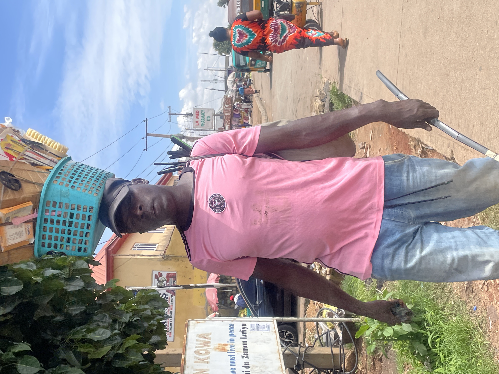 The image captures a man walking down a street in Jos, Nigeria. He is wearing a pink t-shirt and blue jeans. On his head, he carries a blue basket filled with various items, including what appear to be brushes and packaged goods. He is holding a long, thin object in his right hand. In the background, there are buildings, a sign that reads "LA MED PHARMACY LTD," several people, and a tricycle. The street is paved, and there is vegetation on the side. The sky is blue with scattered clouds.