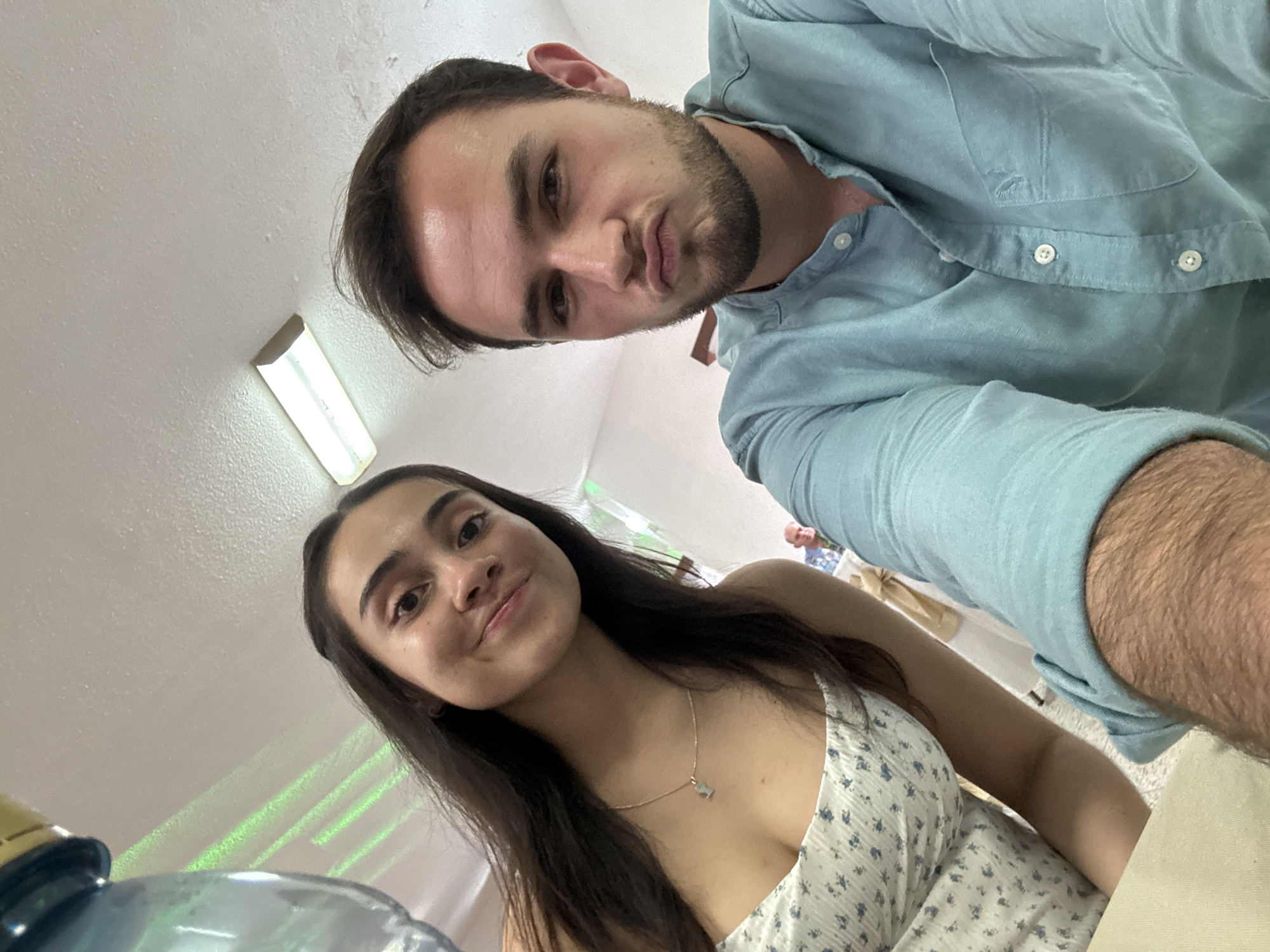 Two individuals, a man and a woman, are in close proximity, likely taking a selfie. The man is wearing a light blue button-down shirt. The woman is wearing a white floral print dress and a necklace. They appear to be indoors, possibly at a gathering or event, as suggested by the presence of other people and objects in the background. The location is Chipilo de Francisco Javier Mina (Chipilo), Mexico. Fluorescent lights are visible on the ceiling. A water dispenser is partially visible in the lower-left corner.