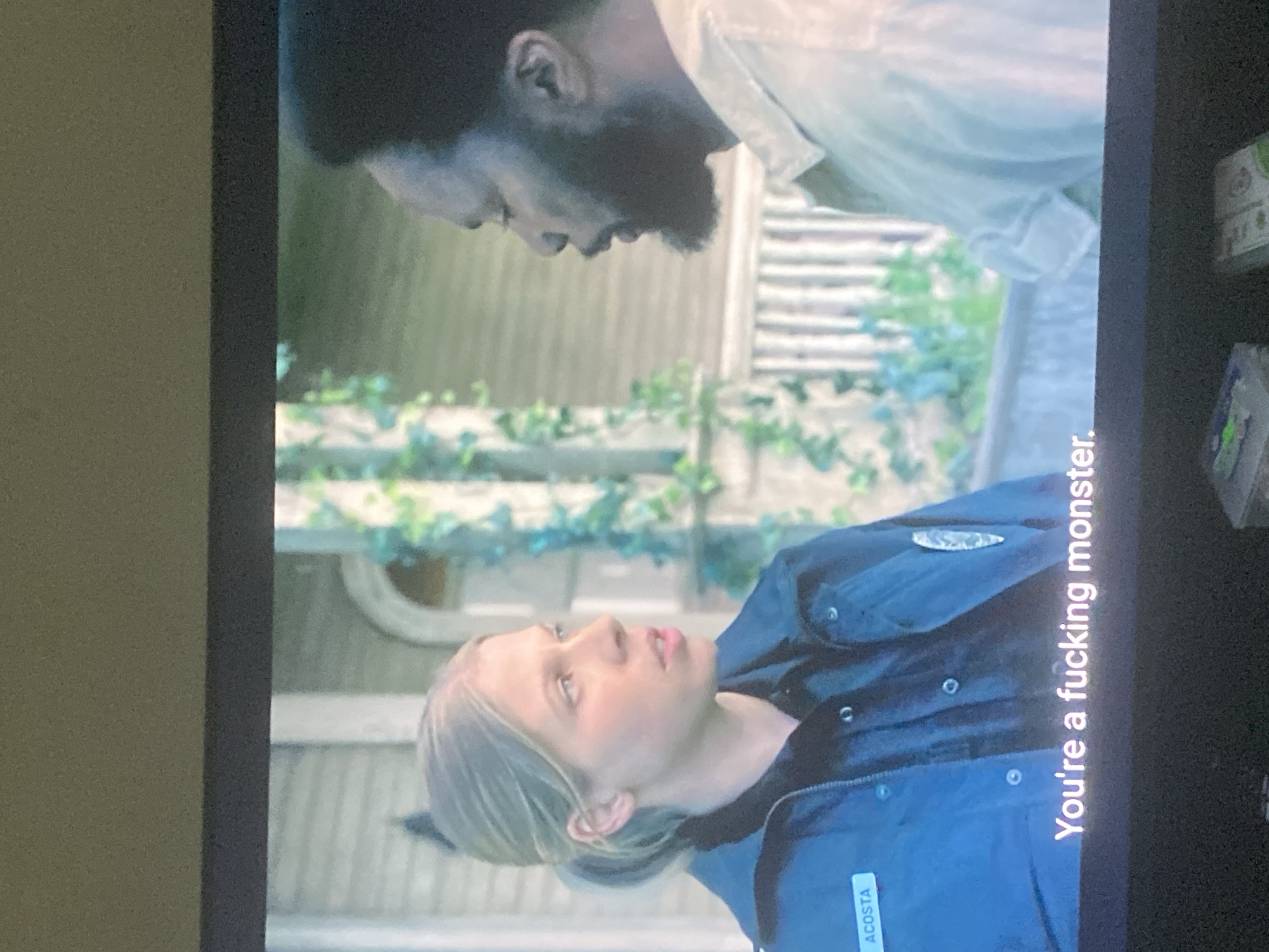 A screen displays a scene featuring two individuals. One is a woman with blonde hair, wearing a blue jacket with the name "ACOSTA" visible on it. She is looking up with an expression of accusation. Text on the screen reads "You're a fucking monster." Above her is a man with dark skin and a beard, leaning down towards her. The background shows a building covered in green vines. The scene is displayed on a television or computer screen, with the frame of the screen visible. There is no evidence to identify the location as Abuja, Nigeria.