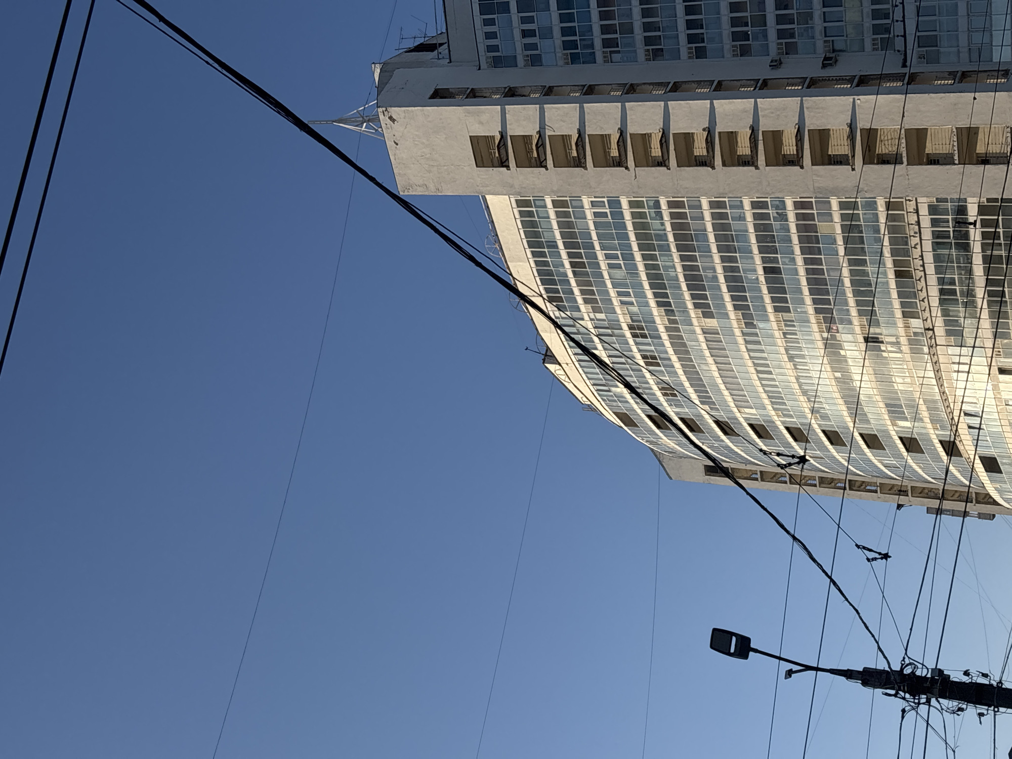 A low-angle shot captures a building with a grid of windows against a clear blue sky. Power lines traverse the scene, intersecting the building and sky. A streetlight is positioned in the lower right corner, connected to the power lines. This is the building of the Sberbank of Russia in Perm, Russia.