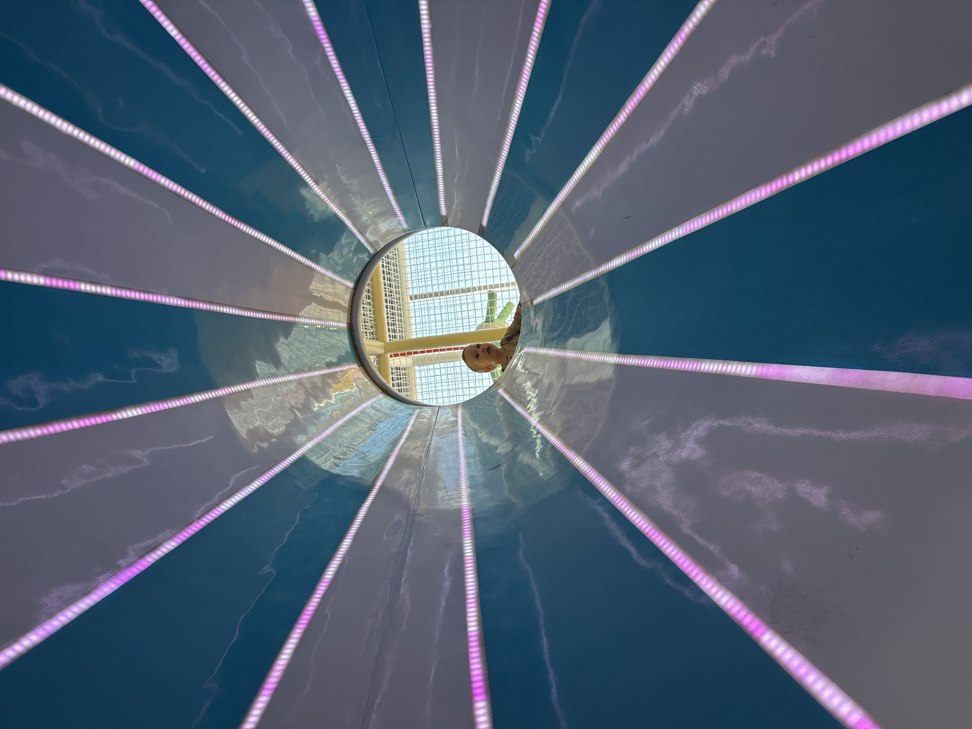 A low-angle shot inside a tunnel with alternating blue and white panels illuminated by vertical rows of pink LED lights converging towards a circular opening. A child's face is visible through the opening. The background visible through the opening suggests an indoor playground or recreational area. The location is likely Perm, Russia, based on metadata or other associated information.