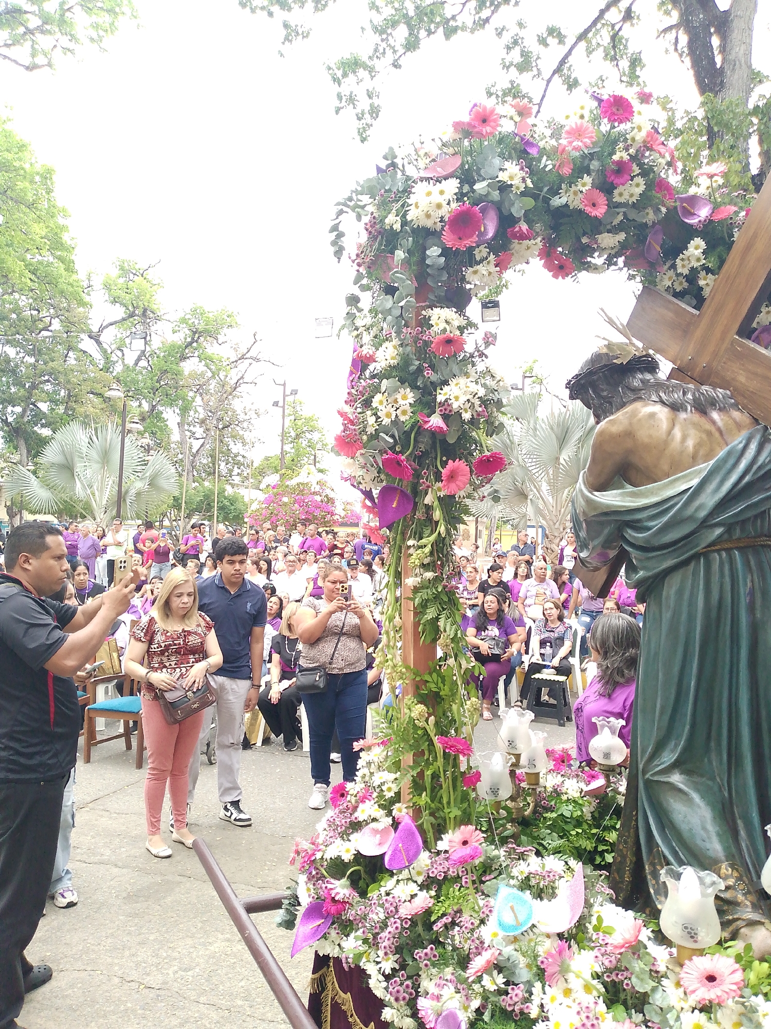 A crowd of people gathers around a statue of Jesus carrying the cross, located in Tinaquillo, Venezuela. Most of the individuals in the crowd are wearing purple shirts. Many are taking pictures with their mobile phones. The statue of Jesus is adorned with an arch of flowers. There are trees and buildings in the background.