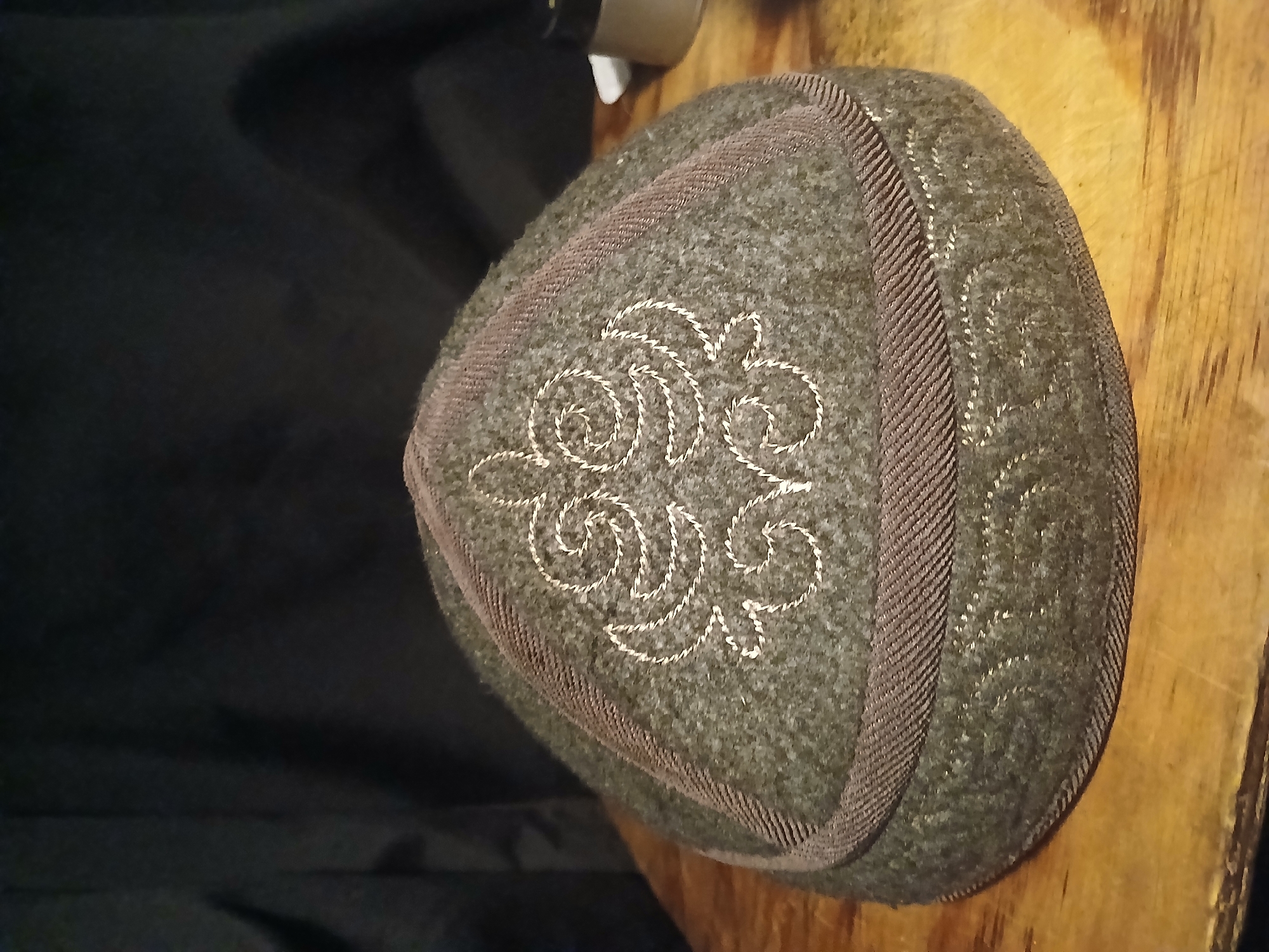 A close-up, eye-level, medium shot captures a felt skullcap with decorative embroidery. The cap is a muted olive green color and sits on a light brown wooden surface. A band of darker brown braid encircles the base of the cap, and a similar braid trims the edge of the cap. White embroidery adorns the top of the cap with a swirling, symmetrical pattern, and another band of white embroidery with a repeating wave-like design encircles the lower portion of the cap. The background is dark and out of focus, suggesting a dimly lit interior. There are no people or animals visible in the image. The scene does not provide any information to suggest a location in Douai, France.