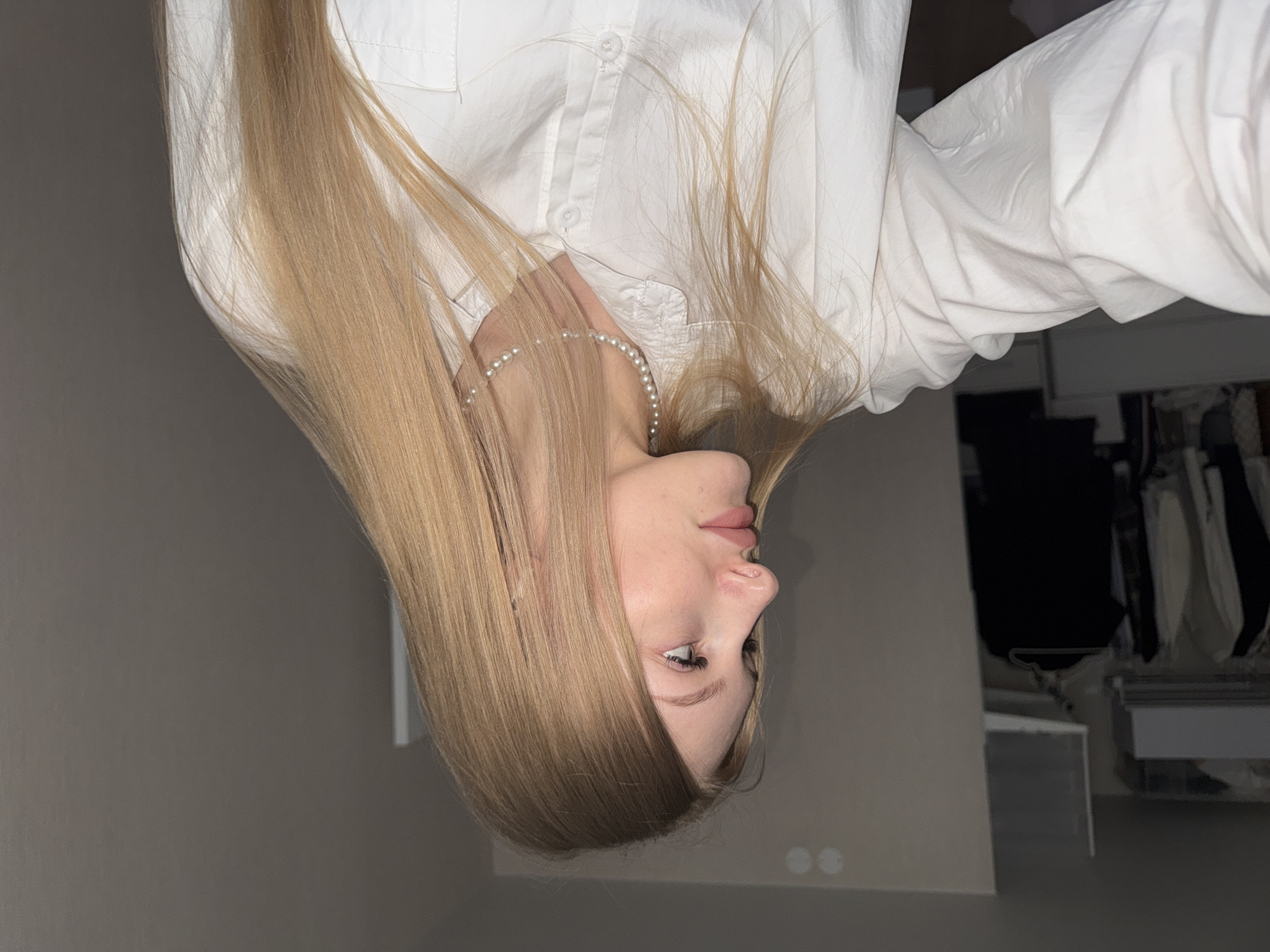 A young woman is depicted indoors, wearing a white button-down shirt and a pearl necklace. Her long blonde hair is cascading. The background suggests an interior setting, possibly a bedroom, with a closet visible. No evidence indicates the location in Perm, Russia.