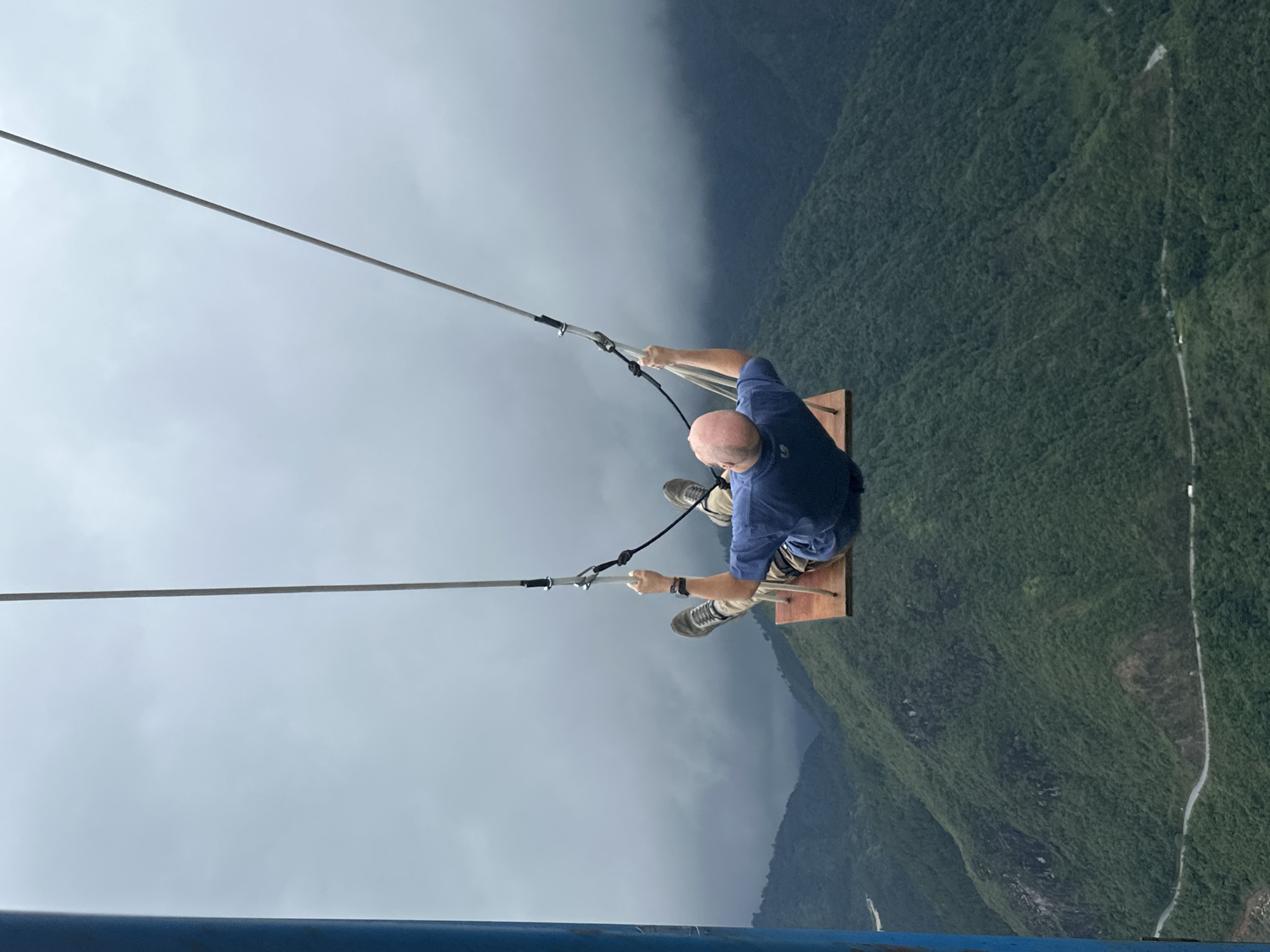 A man is on a swing that is precariously located over the edge of a mountain in Binh Lu, Vietnam. He is wearing a blue shirt, khaki pants, and sneakers. He is holding onto the swing's ropes and appears to be enjoying the view. Below him are lush green mountains and a road. The sky is overcast with dark clouds. A blue railing is visible in the lower left corner of the image, suggesting the swing is situated on some kind of platform.