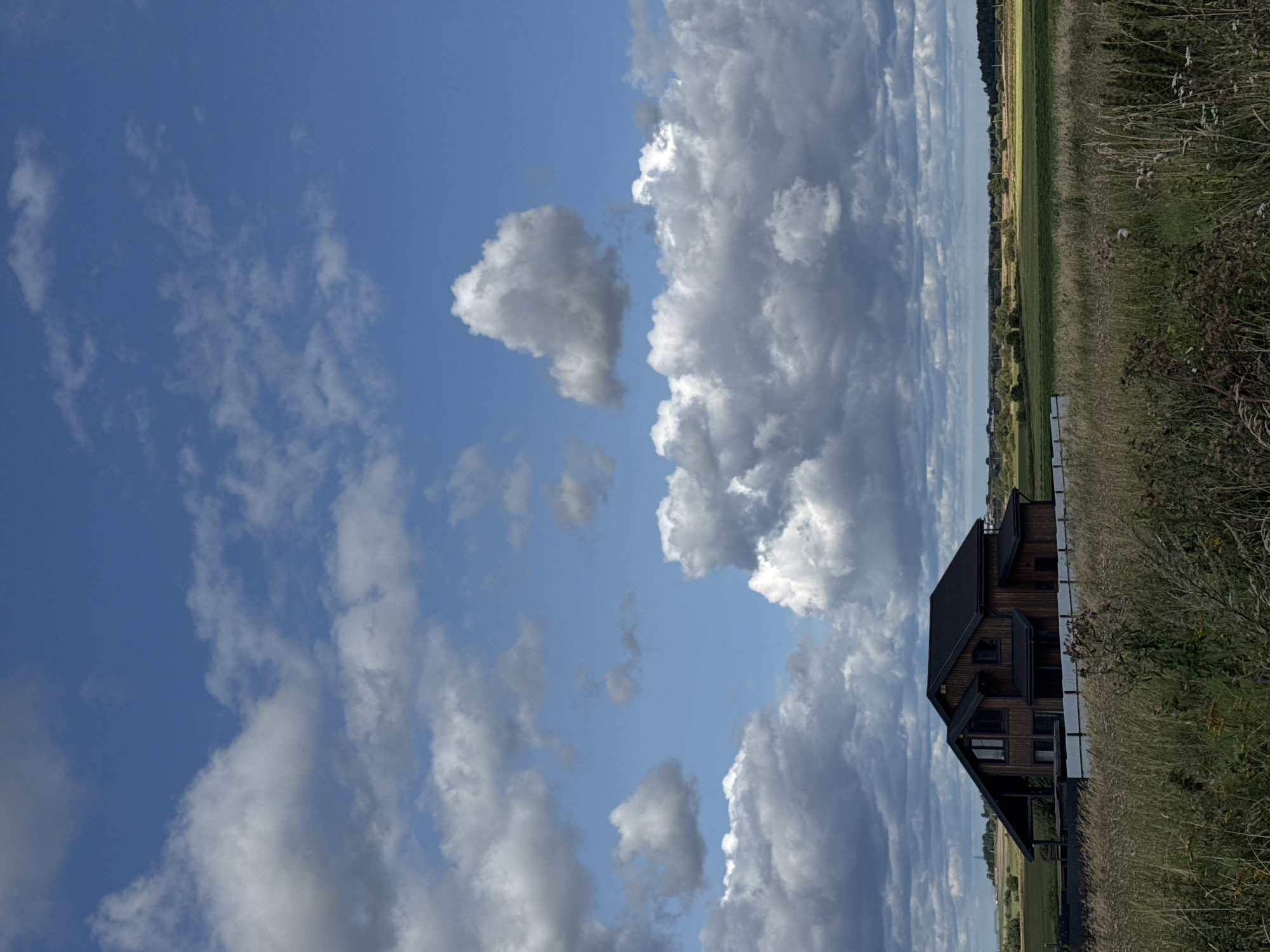 The image depicts a modern wooden house with a dark roof, situated in a rural landscape in Kempelevo, Russia. The house is located behind a white fence and surrounded by tall, dry grass in the foreground. Behind the house, there are green fields and a line of trees in the distance. The sky is filled with dramatic, puffy white and gray clouds against a bright blue backdrop. The overall scene suggests a serene and natural environment.