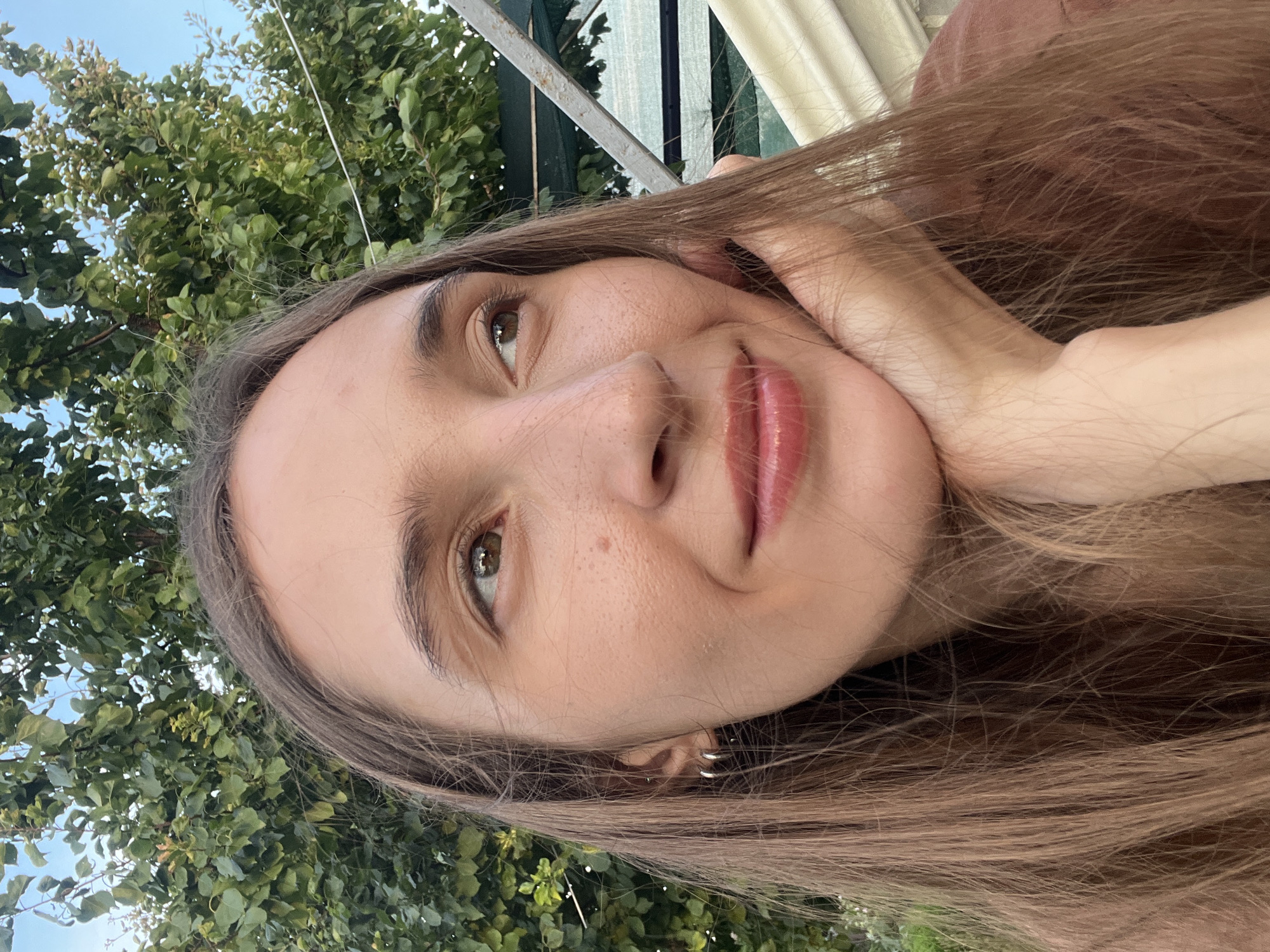 A medium close-up shot shows a young woman resting her chin on her hand, looking to her right, with long, light brown hair framing her face. She is wearing a reddish-brown top and small silver hoop earrings in her left ear. The background is softly blurred, featuring lush green foliage from a tree and hints of a light blue sky. A metal structure with a green tarp is visible behind her on the right. The lighting suggests an outdoor setting, likely during the day.