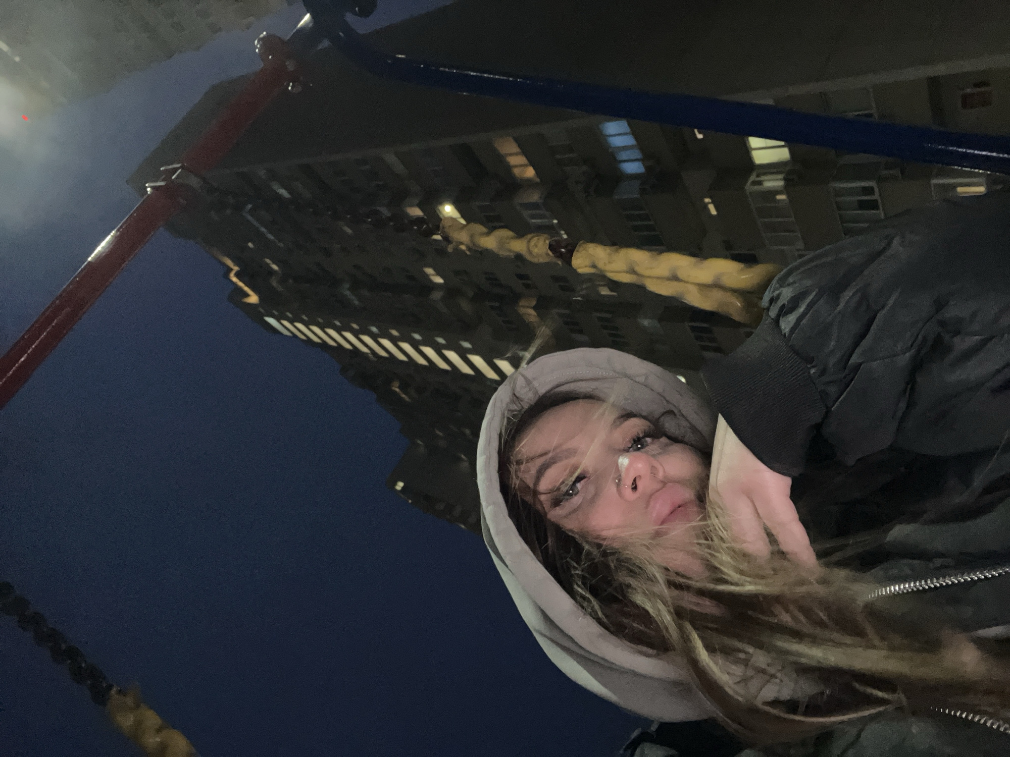 This tilted, low-angle image captures a young woman outdoors at night in Krasnodar, Russia. The central subject is seen looking directly at the camera, making a slight pouting expression. She has long, light brown hair, a visible nose piercing, and makeup emphasizing her eyes. She is wearing a light grey hooded sweatshirt with the hood up, underneath a dark, likely black, puffy jacket with a prominent zipper. One of her hands is visible, resting near her hair and face on the right side of the frame.
The setting appears to be an urban or residential area. In the background, a large, multi-story building, possibly an apartment complex, rises against the dark night sky. Many of its windows are illuminated, confirming the time of day as night. In the immediate foreground and partially framing the woman, there are structural elements that suggest a playground swing set, including red and blue metal bars and a thick, yellow-wrapped chain.
The activity taking place is a casual portrait or selfie. The woman's attire, particularly the puffy jacket, implies cool or cold weather. There is no visible text in the image.