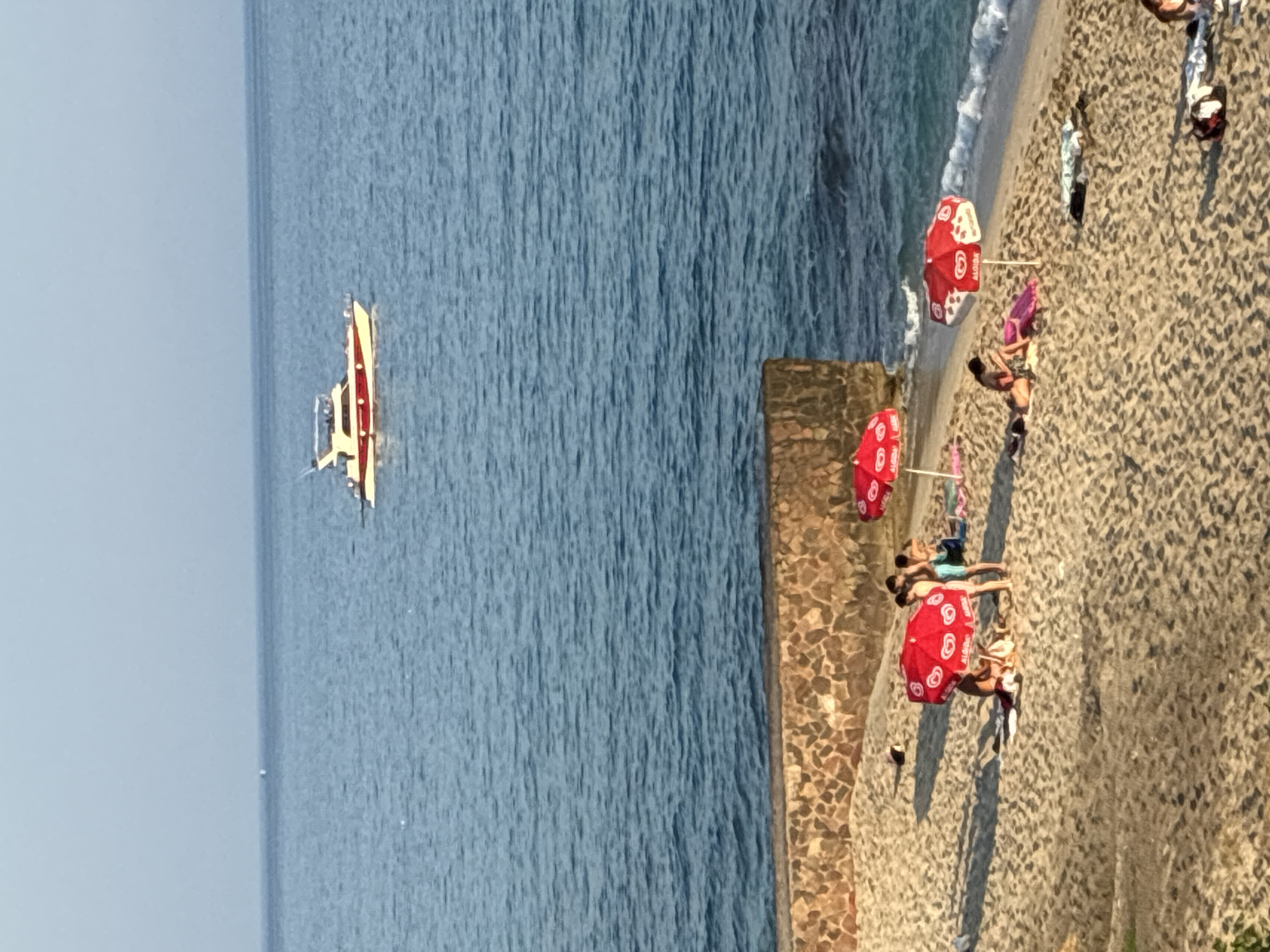 In Alanya, Turkey, a scenic view captures a serene beach scene with the tranquil blue sea stretching to the horizon. A white and red motorboat cruises on the water, while the sandy shore is dotted with beachgoers enjoying the sun. Red umbrellas, some bearing the Algida ice cream logo, provide shade for people lounging on the sand. A stone wall separates the beach from a grassy area, adding a rustic touch to the landscape. The gentle waves of the sea kiss the shore, creating a peaceful and idyllic atmosphere.