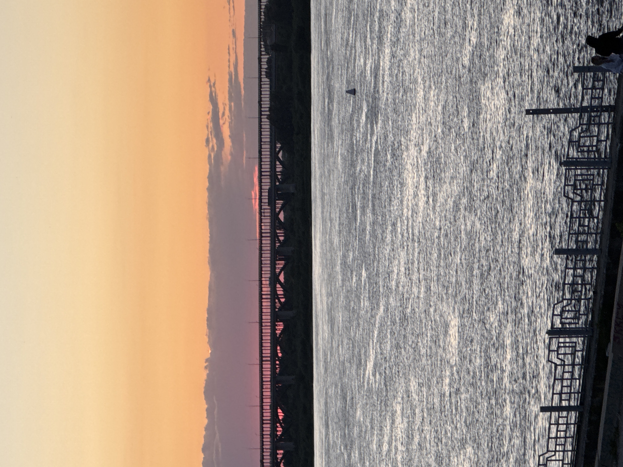 The image shows a sunset over a body of water, likely a river, as evidenced by the presence of a large bridge. The sky transitions from a pale orange at the horizon to a soft yellow higher up, with faint clouds visible. A portion of the bridge is silhouetted against the colorful sky, revealing its structural details. On the right side of the image, the water is shimmering, reflecting the light of the setting sun. A buoy can be seen in the water. Along the edge of the water, there is a walkway with railings and steps leading down to the water. A person is partially visible on this walkway, silhouetted against the bright water. The overall mood is peaceful and serene, capturing the beauty of a sunset by the water in Nizhny Novgorod, Russia. The time of day is clearly sunset.