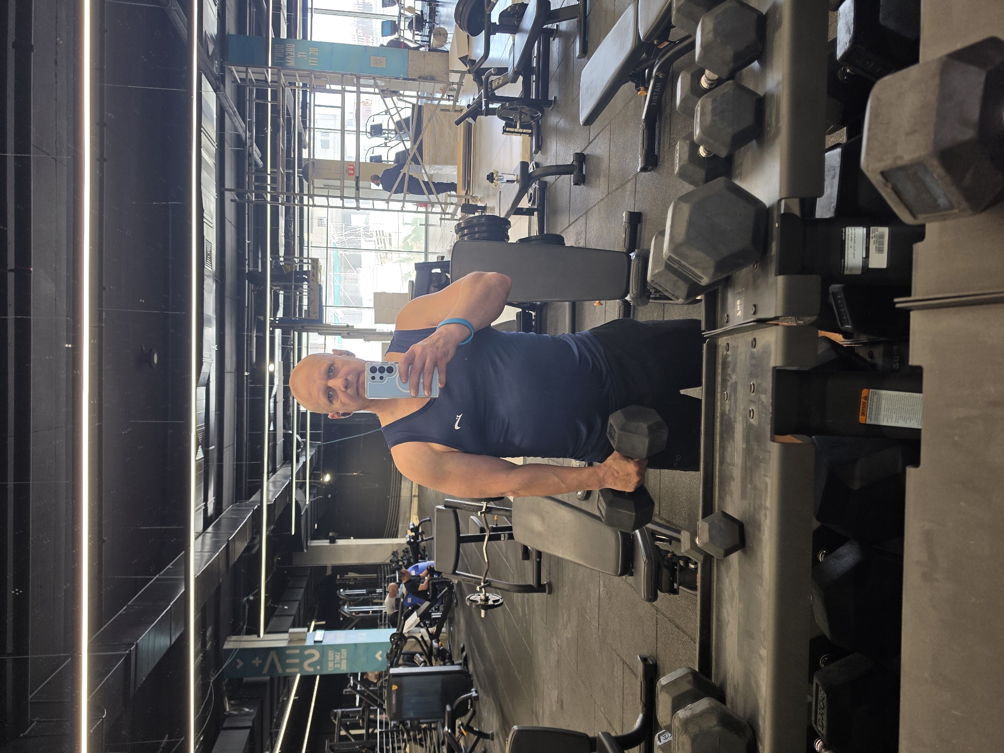 Here is a detailed description of the image:
The image depicts an individual in what appears to be a fitness center or gym. The person, a bald man wearing a navy blue sleeveless shirt and black shorts, is positioned in front of a mirror, using a smartphone to take a selfie. He holds a dumbbell in his right hand, suggesting he is in the middle of a workout.
The environment is filled with various exercise equipment, including dumbbell racks filled with weights, weight benches, and other gym machinery. The color scheme is predominantly grey and black, with metal and rubber surfaces visible. There are other individuals in the background, also engaged in exercise activities.
Through the windows, there is a view of an urban landscape with buildings, indicating an indoor setting with access to natural light. Given the OCR text in the image (specifically "*+2EV") and the location mentioned in the prompt, it is likely the image was taken in Bat Yam, Israel.