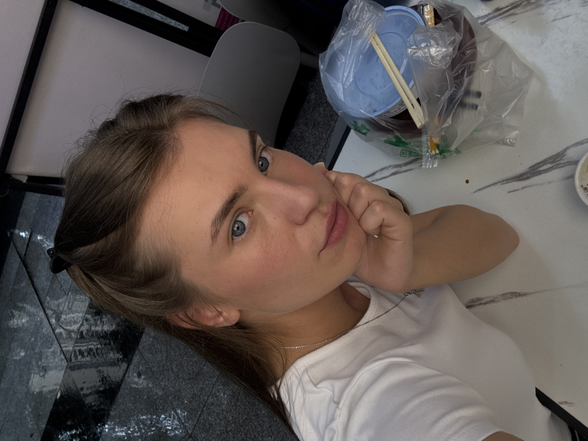 This media file features a young woman in a close-up selfie, oriented vertically. She has fair skin, light brown hair, and blue eyes. Her gaze is directed towards the camera, and her left hand is resting on her cheek. She is wearing a white t-shirt and a delicate silver necklace.
The background suggests an indoor setting, possibly a restaurant or cafe, indicated by a marbled tabletop and a plastic bag containing food items, including chopsticks and what appears to be a blue container. A portion of a grey chair is also visible. The lighting appears to be indoor ambient light, suggesting it could be daytime or evening.
The woman's expression is somewhat pensive or thoughtful. The overall mood is casual and personal, typical of a selfie.
There is no visible text in this media file. The location context provided is Haizhu District, China.